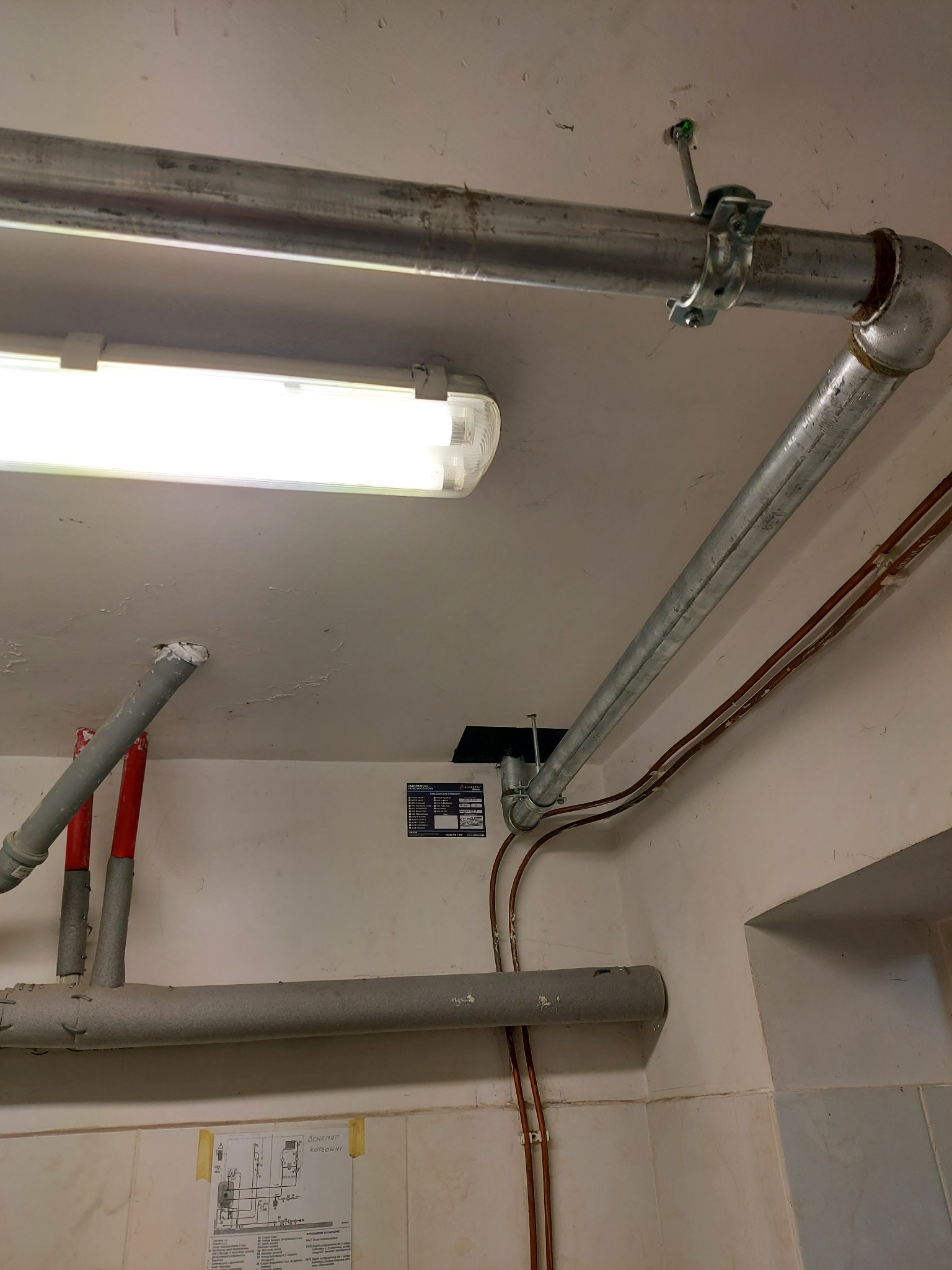 Metalowe rury wentylacyjne i instalacyjne pod sufitem, widoczne mocowania, izolacja termiczna, schemat kotłowni przyklejony do ściany.