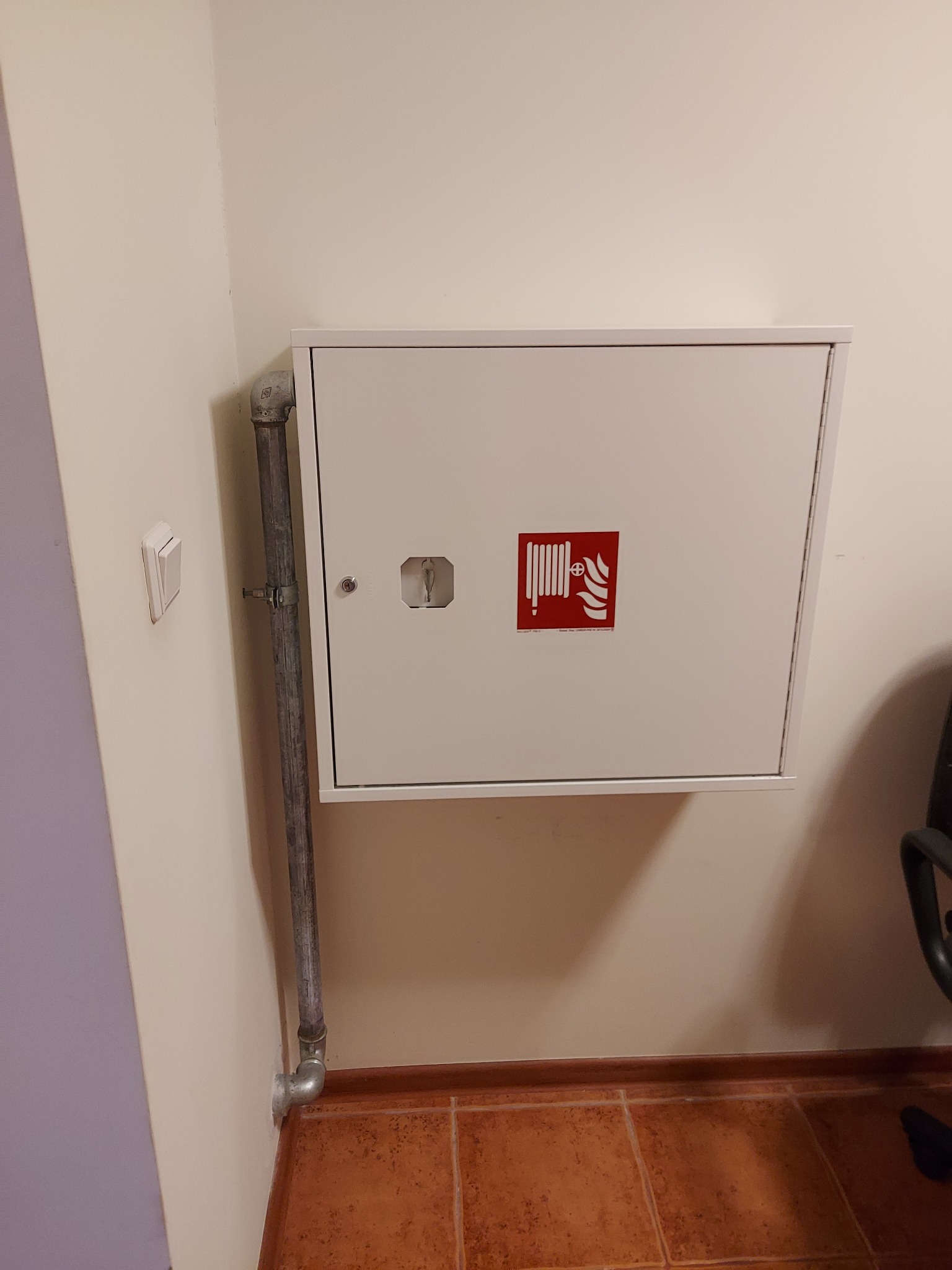 Biała szafka hydrantowa z czerwoną ikoną węża pożarniczego, zamontowana na ścianie obok pionowej rury instalacyjnej w pomieszczeniu z brązową terakotą.