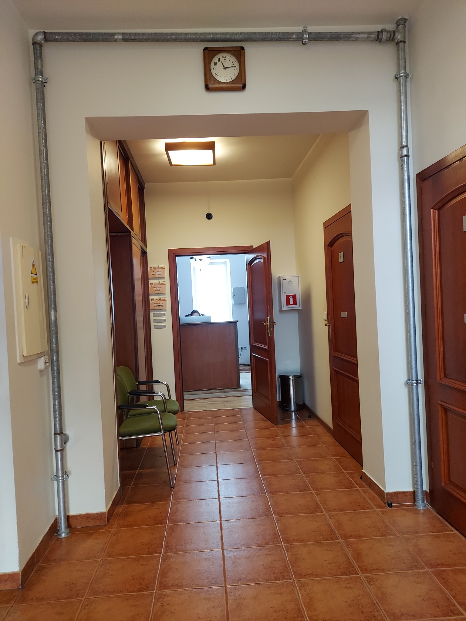Wnętrze biura z widoczną instalacją rur wentylacyjnych pod sufitem, brązowe płytki na podłodze, krzesła dla klientów i zegar na ścianie.
