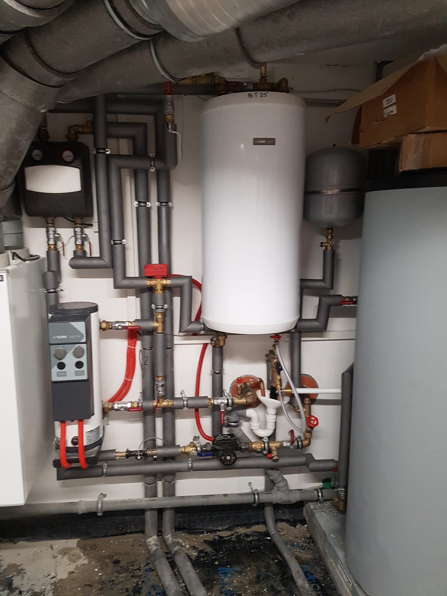 Złożona instalacja grzewcza z białym zasobnikiem ciepłej wody, szarymi rurami izolowanymi, zaworami i osprzętem, widoczna w pomieszczeniu technicznym.