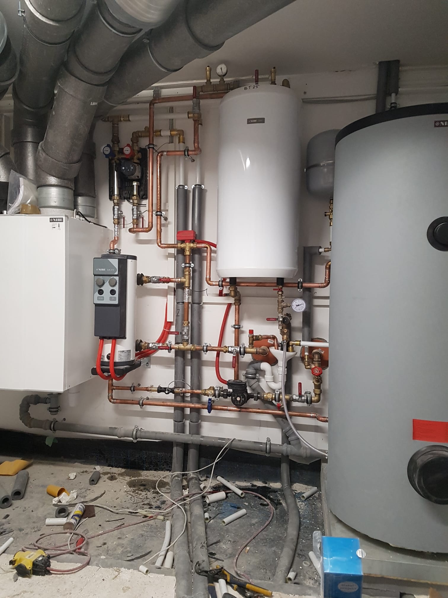 Instalacja grzewcza w kotłowni z dwoma zbiornikami, licznymi rurami miedzianymi i szarymi, widoczny manometr, na podłodze resztki materiałów instalacyjnych.