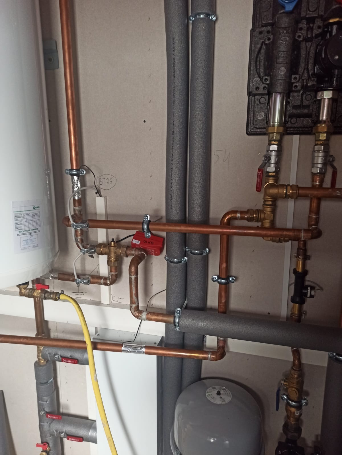 Instalacja grzewcza z miedzianymi rurami, zaworami i izolowanymi przewodami, widoczne elementy kotła, zbiornik wyrównawczy i okablowanie, na ścianie notatki ołówkiem.