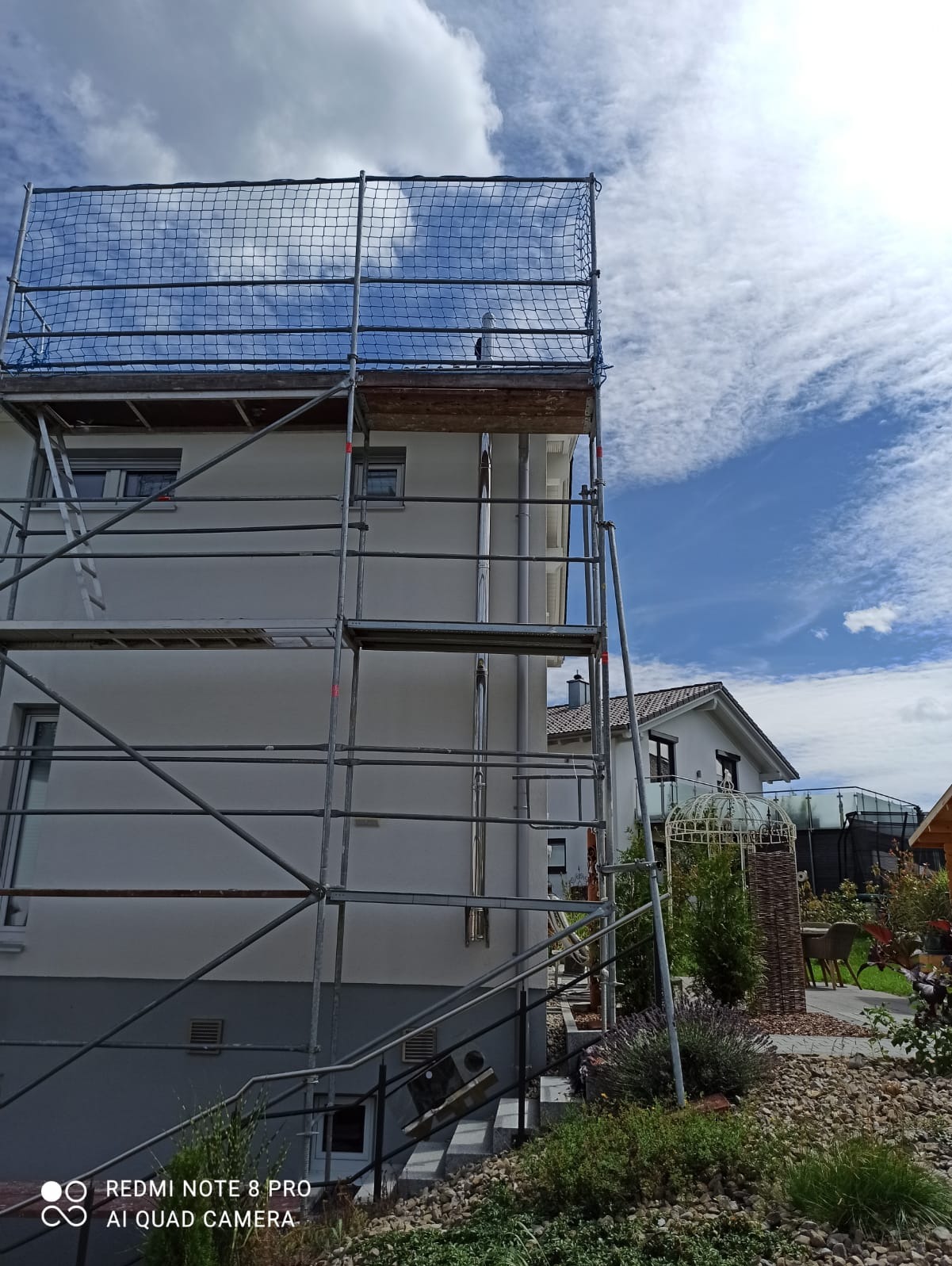 Elewacja domu jednorodzinnego w trakcie prac wykończeniowych, osłonięta rusztowaniem z siatką zabezpieczającą, widok z poziomu ogrodu.