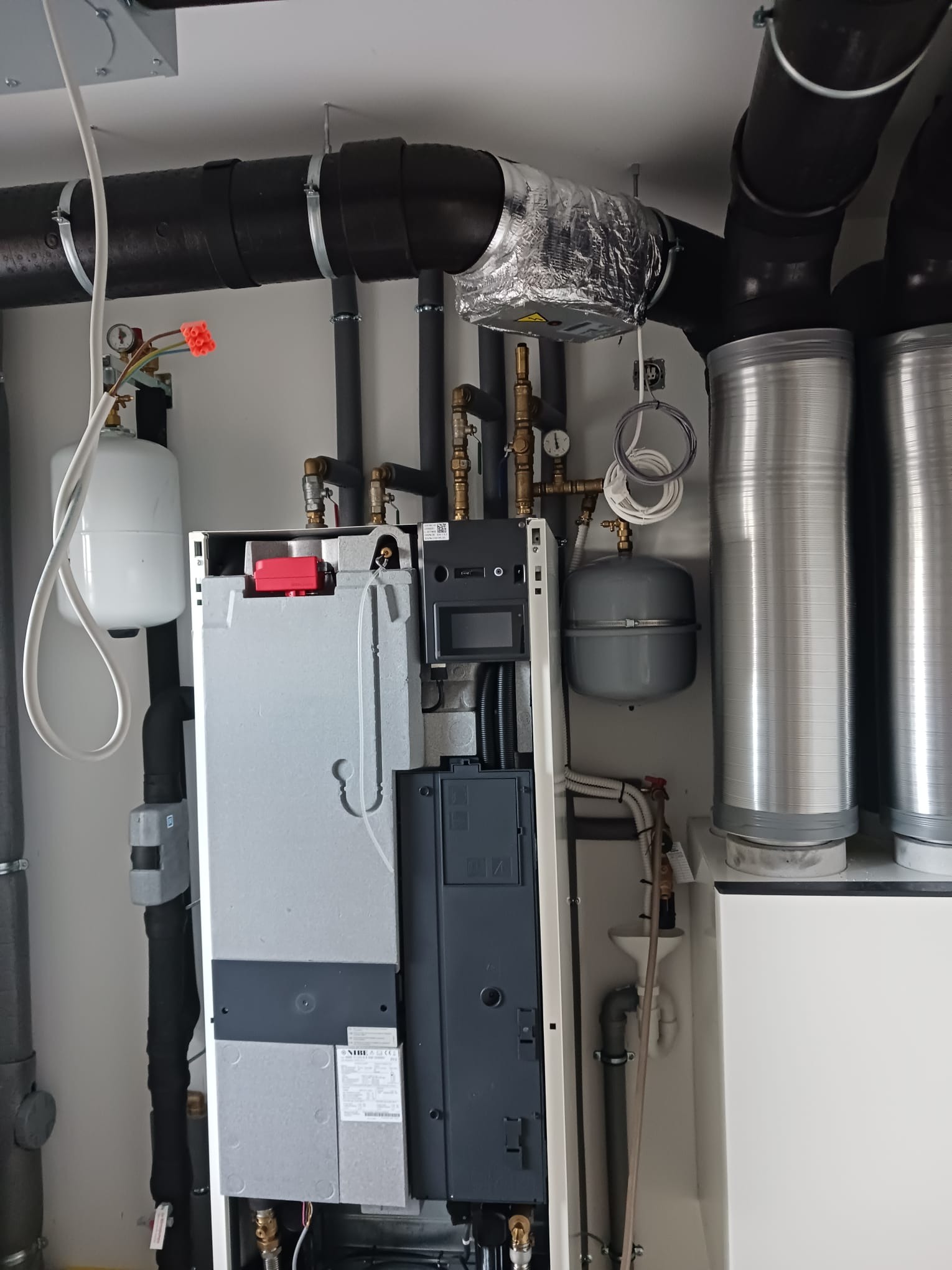 Nowoczesny piec centralnego ogrzewania NIBE z rozbudowaną instalacją rur, zaworów i zbiorników wyrównawczych, widoczne metalowe kominy spalinowe.