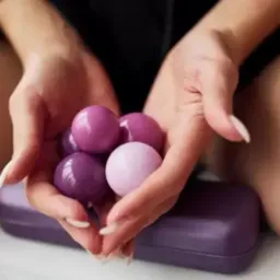 Dłonie kobiety z długimi, białymi paznokciami trzymają garść liliowych i fioletowych, błyszczących kul, na tle liliowego pudełka.