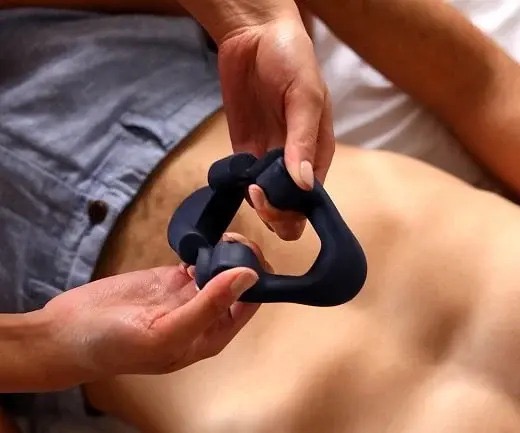 Dwie dłonie trzymają futurystyczny, ergonomiczny gadżet w kolorze ciemnego grafitu, przypominający trójkąt z zaokrąglonymi wypustkami, na tle fragmentu ciała mężczyzny w szortach, leżącego...