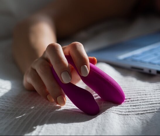 Dłoń kobiety z beżowym manicure trzyma fioletowy gadżet erotyczny na tle białej pościeli i rozmytego laptopa.