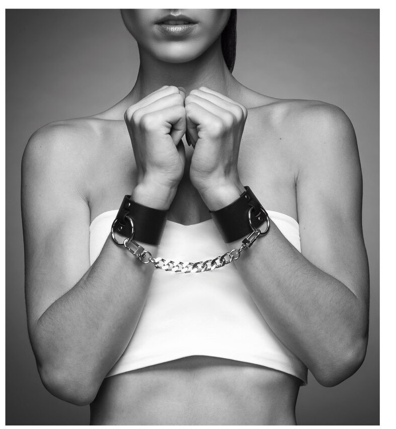 Kobieta w białym topie z czarnymi skórzanymi kajdankami na nadgarstkach połączonych srebrnym łańcuchem, dłonie zaciśnięte w pięści, zbliżenie, czarno-białe zdjęcie.