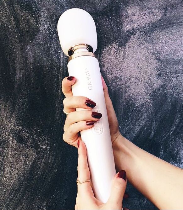 Dłonie kobiety z bordowymi paznokciami trzymają biały masażer elektryczny na tle ciemnoszarej, teksturowanej ściany.