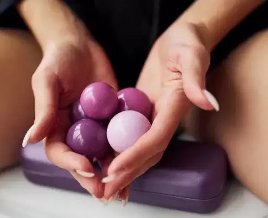 Dłonie kobiety z długimi, białymi paznokciami trzymają garść liliowych i fioletowych, błyszczących kul, na tle liliowego pudełka.