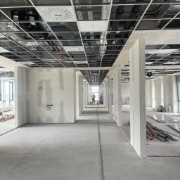 Optima Projekt - Długi korytarz w surowym stanie, ściany częściowo pokryte gładzią, widoczne instalacje pod sufitem, pracownik w oddali.