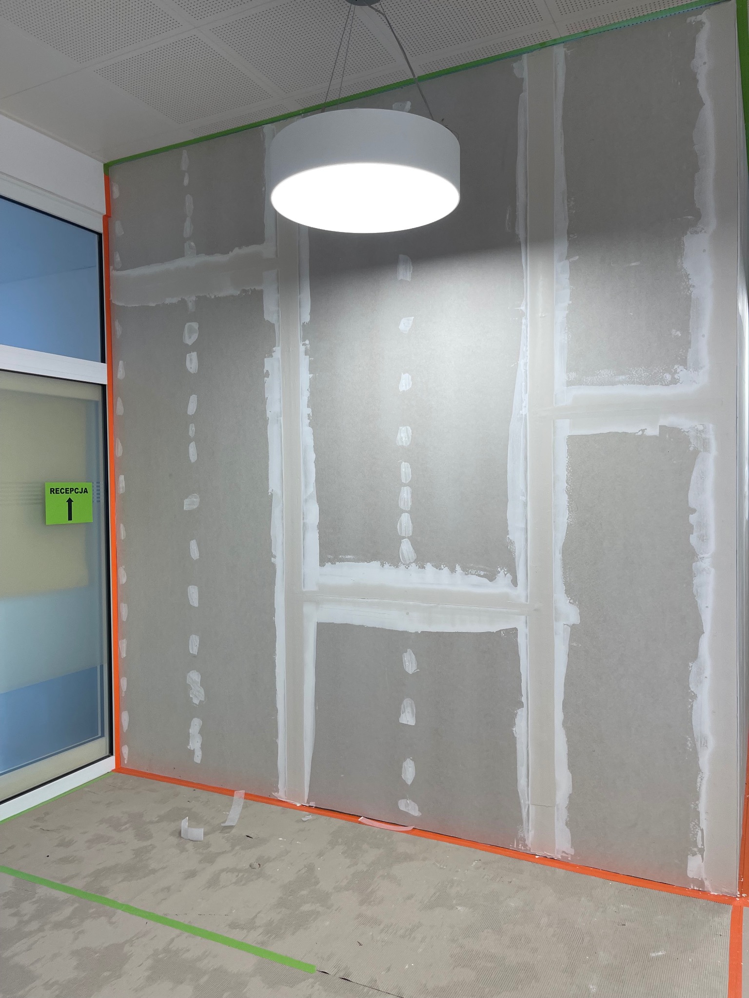 Montaż płyty karton gips na ścianie w pomieszczeniu biurowym, widoczne szpachlowane łączenia, taśma malarska zabezpieczająca narożniki i podłogę, lampa sufitowa w tle, tabliczka 'Recepcja'...
