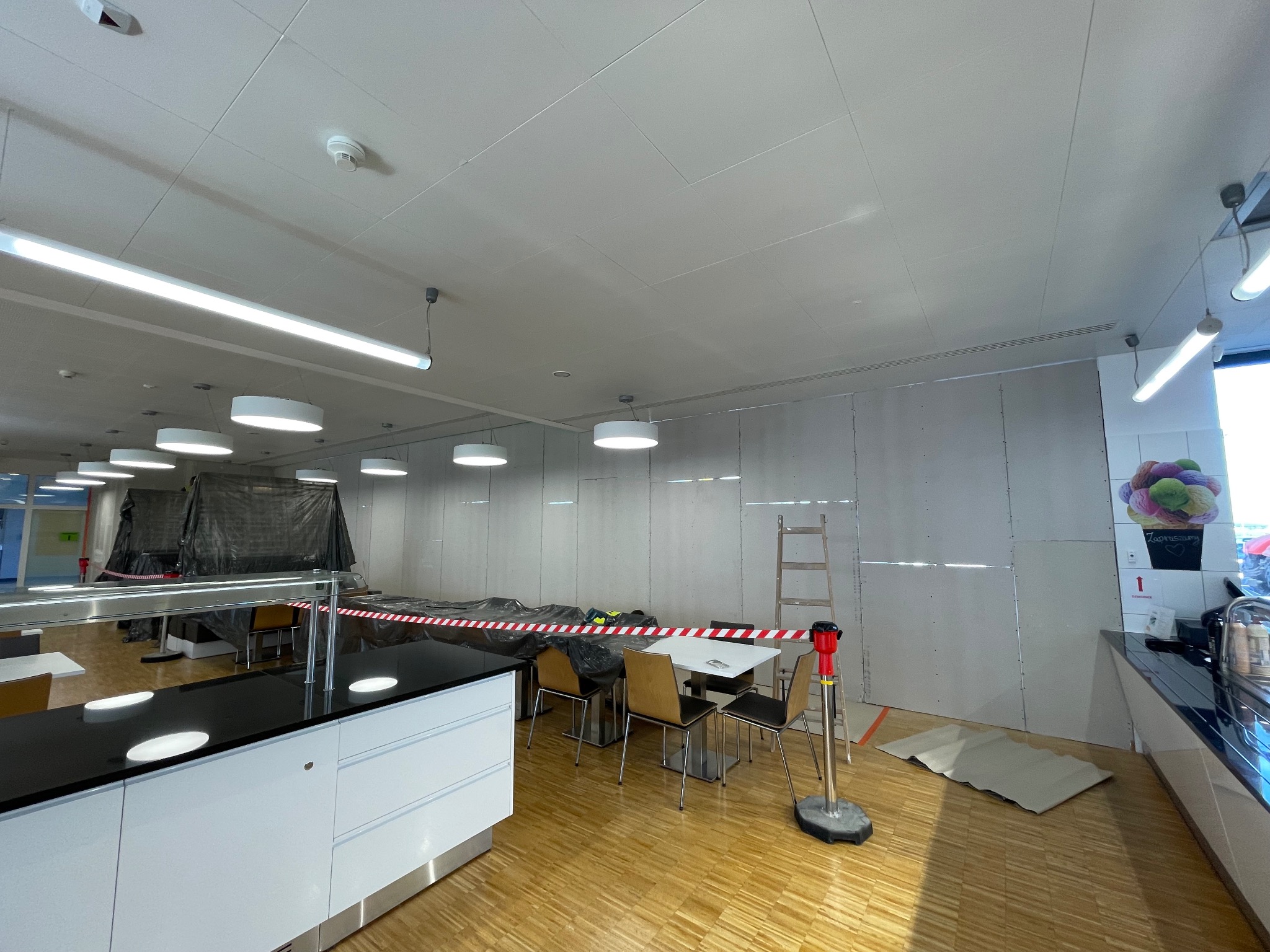 Wnętrze lokalu w trakcie prac wykończeniowych; widoczna ściana z płyt gipsowo-kartonowych, zabezpieczone meble i podłoga, drabina oparta o ścianę.