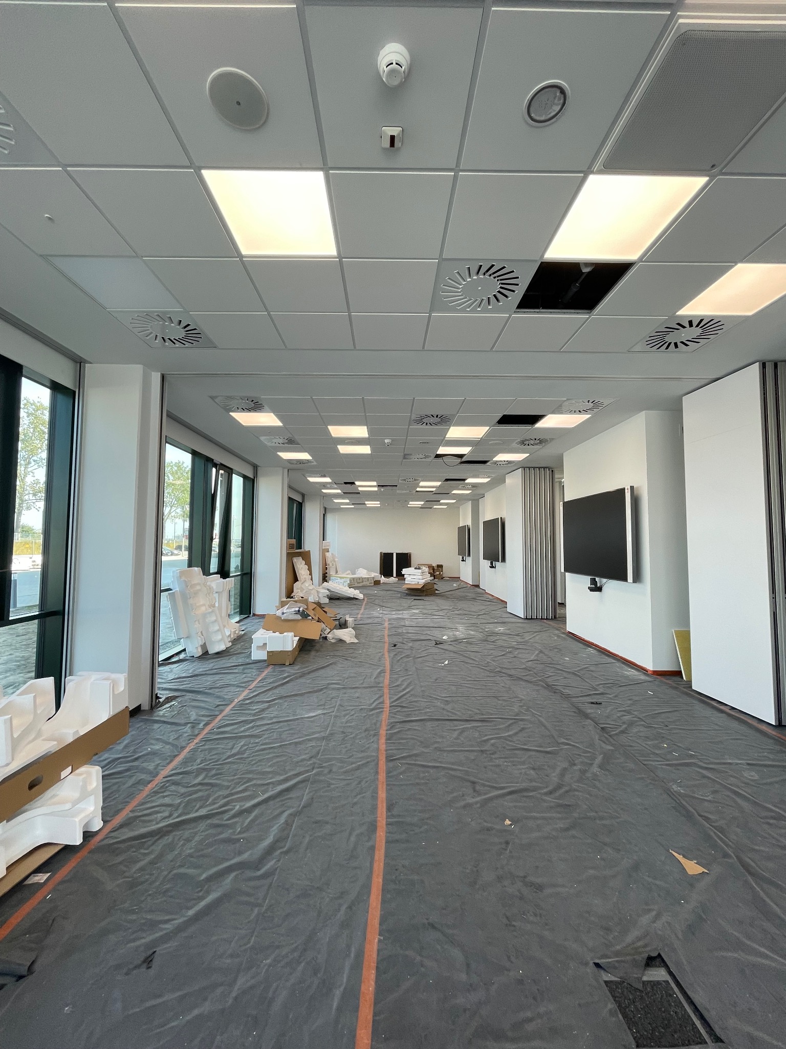 Wnętrze biurowe w trakcie prac wykończeniowych, podłoga zabezpieczona folią, widoczne kartony i elementy montażowe, sufit podwieszany z oświetleniem, duże okna i przesuwane ściany działowe.