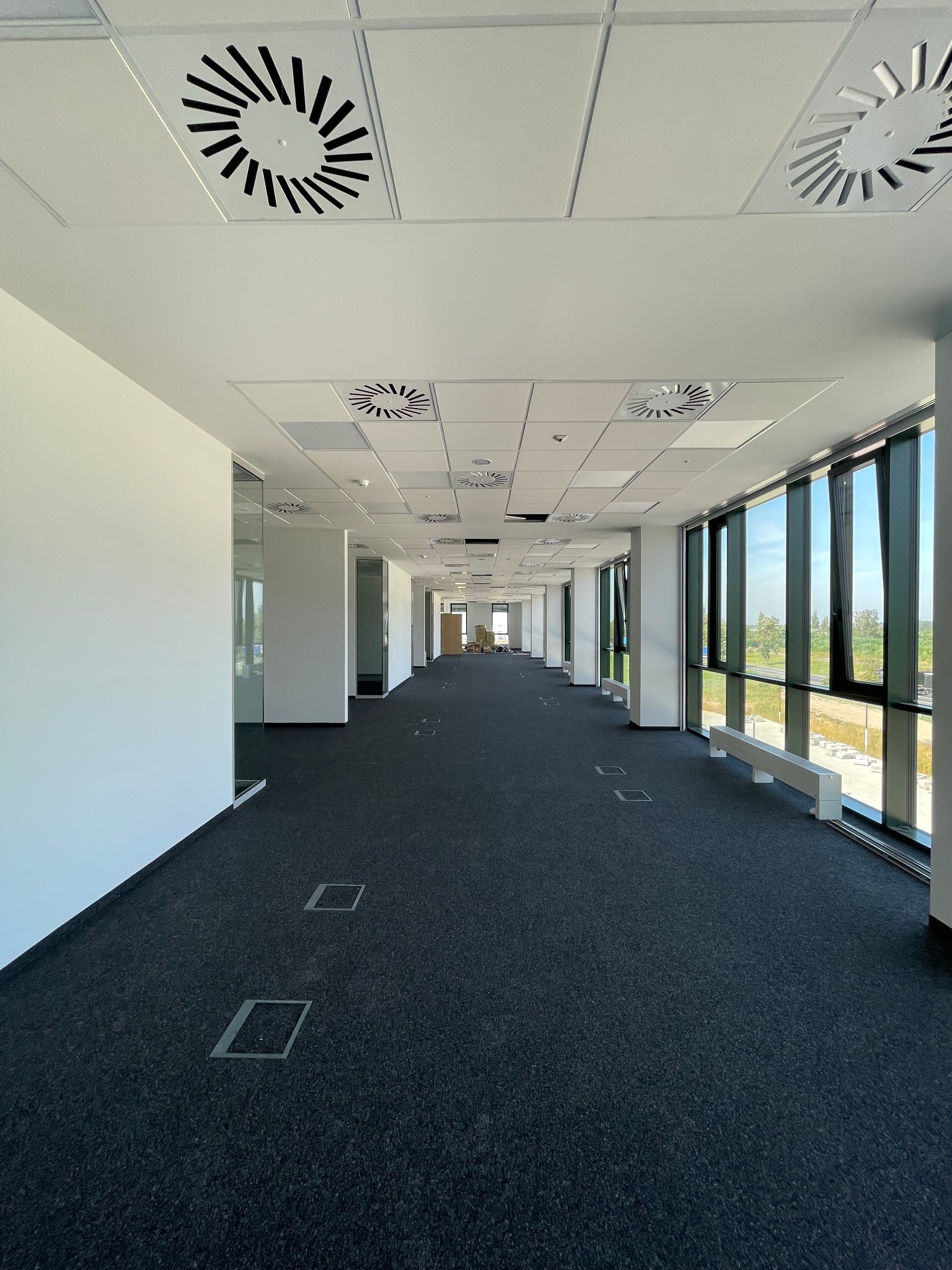 Długi, pusty korytarz biurowy z czarną wykładziną, białymi ścianami i sufitem kasetonowym z okrągłymi kratkami wentylacyjnymi, duże okna po prawej stronie wpuszczają naturalne światło.