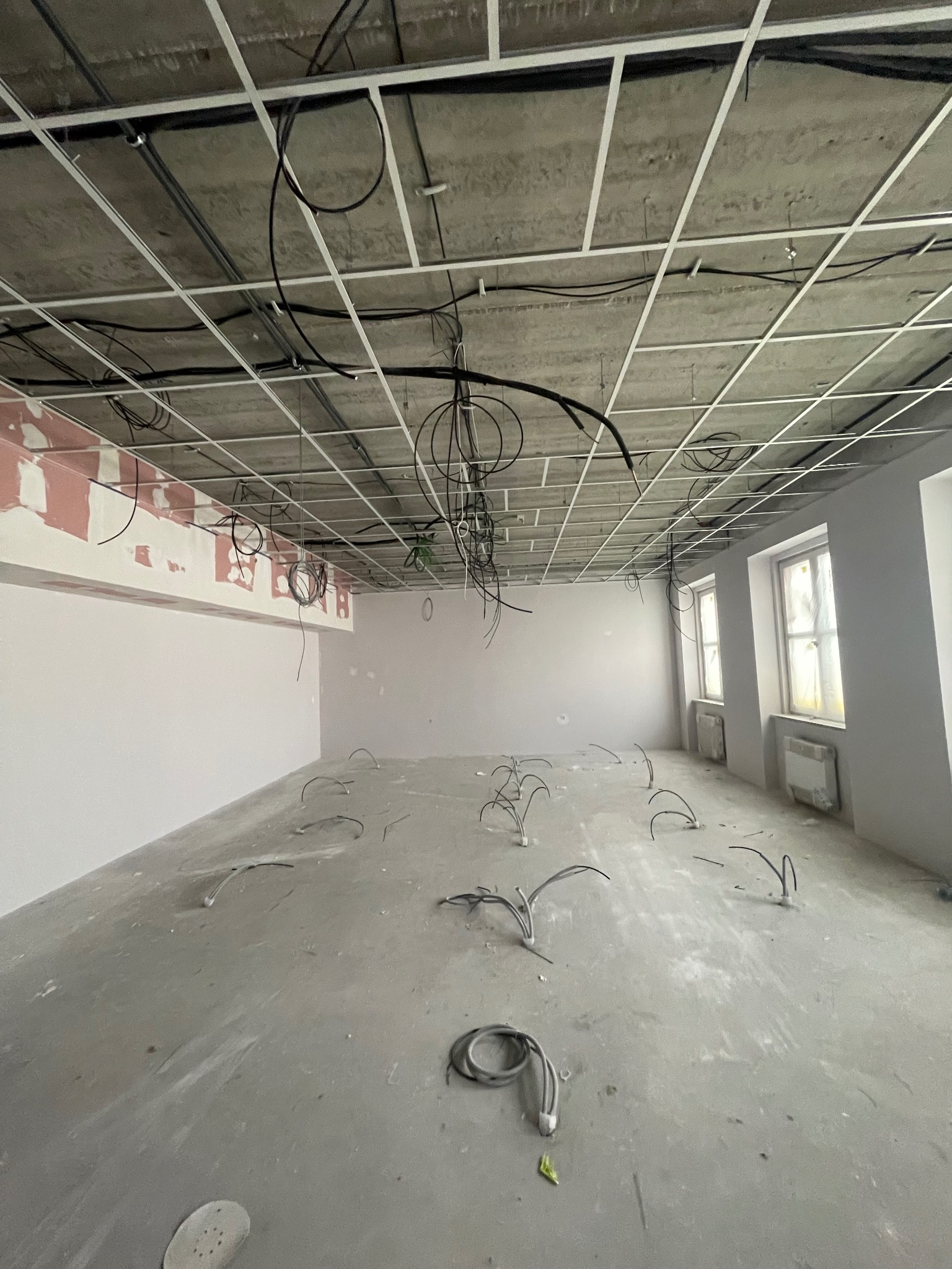 Wnętrze w trakcie prac wykończeniowych: surowy strop z widocznymi przewodami elektrycznymi, ściany przygotowane do malowania, podłoga betonowa z wystającymi kablami, okna zabezpieczone folią.