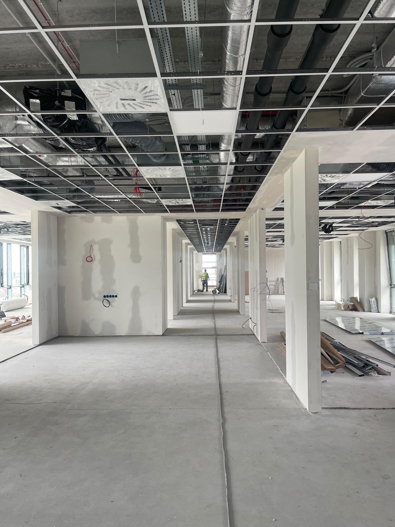Długi korytarz w surowym stanie, ściany częściowo pokryte gładzią, widoczne instalacje pod sufitem, pracownik w oddali.
