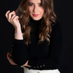 Portret kobiety z brązowymi włosami, w czarnym golfie i białej spódnicy, z delikatnym uśmiechem i błyszczącą bransoletką, na czarnym tle.