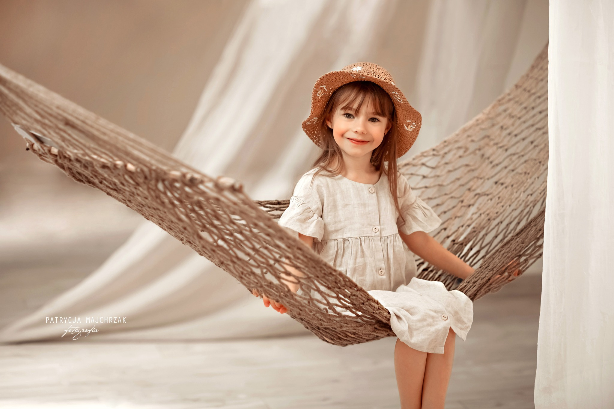 Uśmiechnięta dziewczynka w słomkowym kapeluszu i lnianej sukience siedzi w hamaku w pastelowych barwach na neutralnym tle.