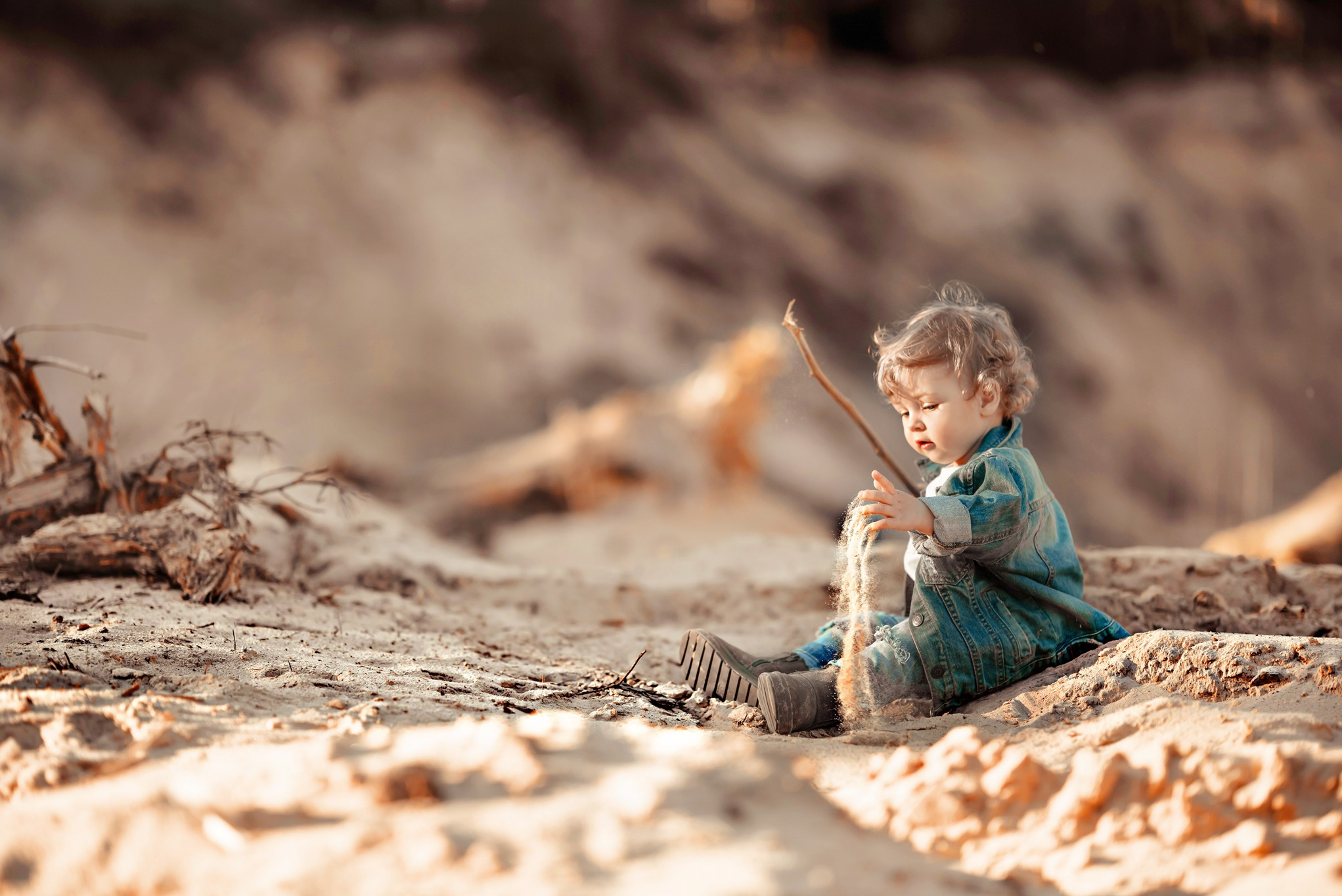Mały chłopiec w dżinsowej kurtce siedzi na piaszczystej plaży, przesypując piasek przez palce w promieniach zachodzącego słońca, w tle rozmyte drzewa i gałęzie.