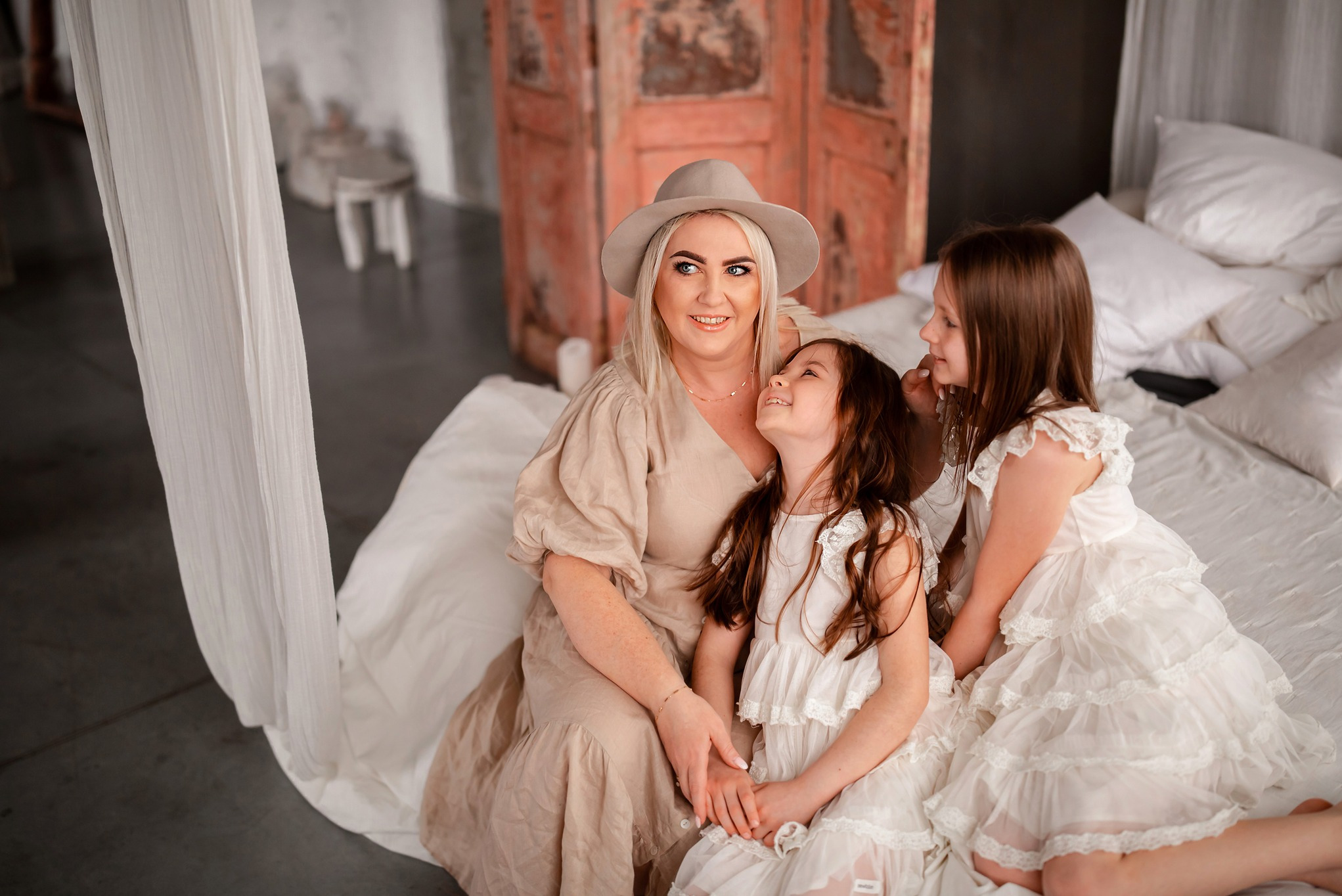 Kobieta w kapeluszu siedzi z dwiema dziewczynkami na łóżku z baldachimem, jasne ubrania, uśmiechnięte twarze, rustykalne tło z drewnianym parawanem.