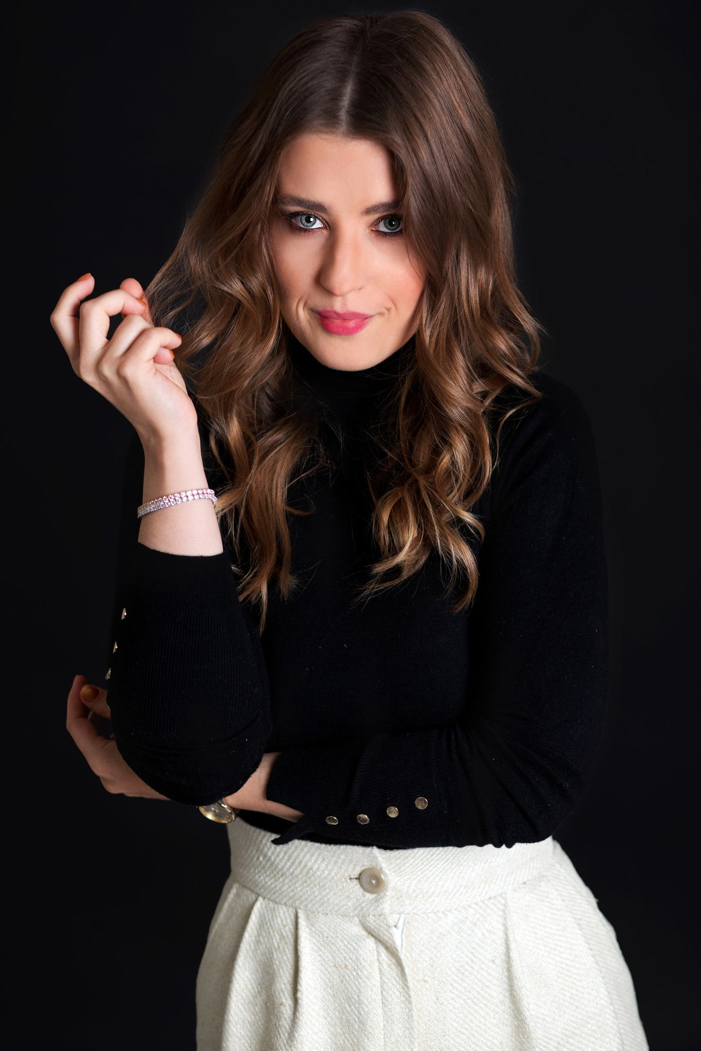 Portret kobiety z brązowymi włosami, w czarnym golfie i białej spódnicy, z delikatnym uśmiechem i błyszczącą bransoletką, na czarnym tle.