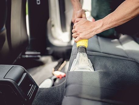 Czyszczenie ciemnej tapicerki samochodowej za pomocą odkurzacza ekstrakcyjnego z przezroczystą dyszą i żółtą rączką, widoczne ślady użycia środka czyszczącego.