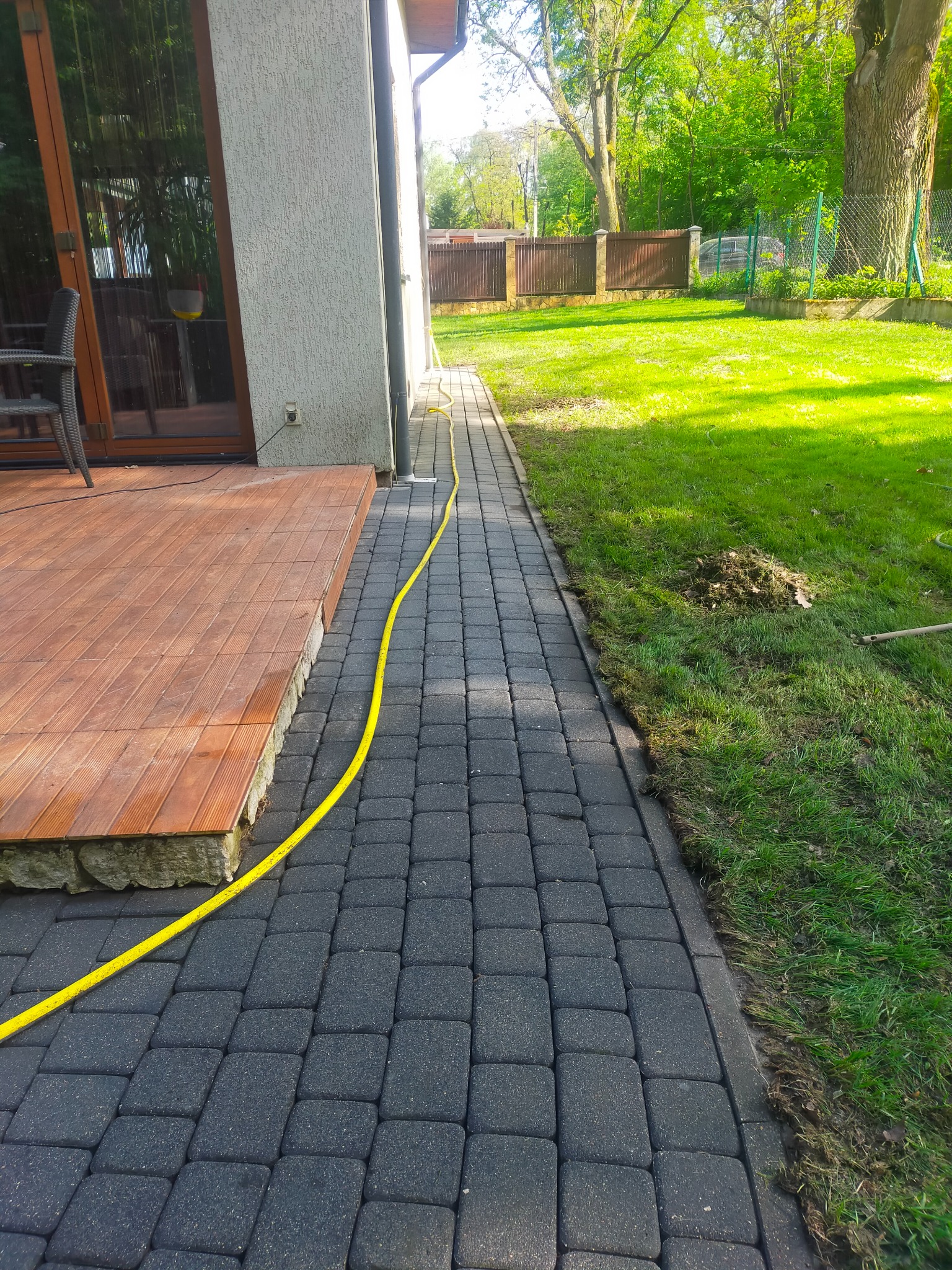Ciemnoszara kostka brukowa ułożona wzdłuż drewnianego tarasu i trawnika, z żółtym wężem ogrodowym leżącym na jej powierzchni.