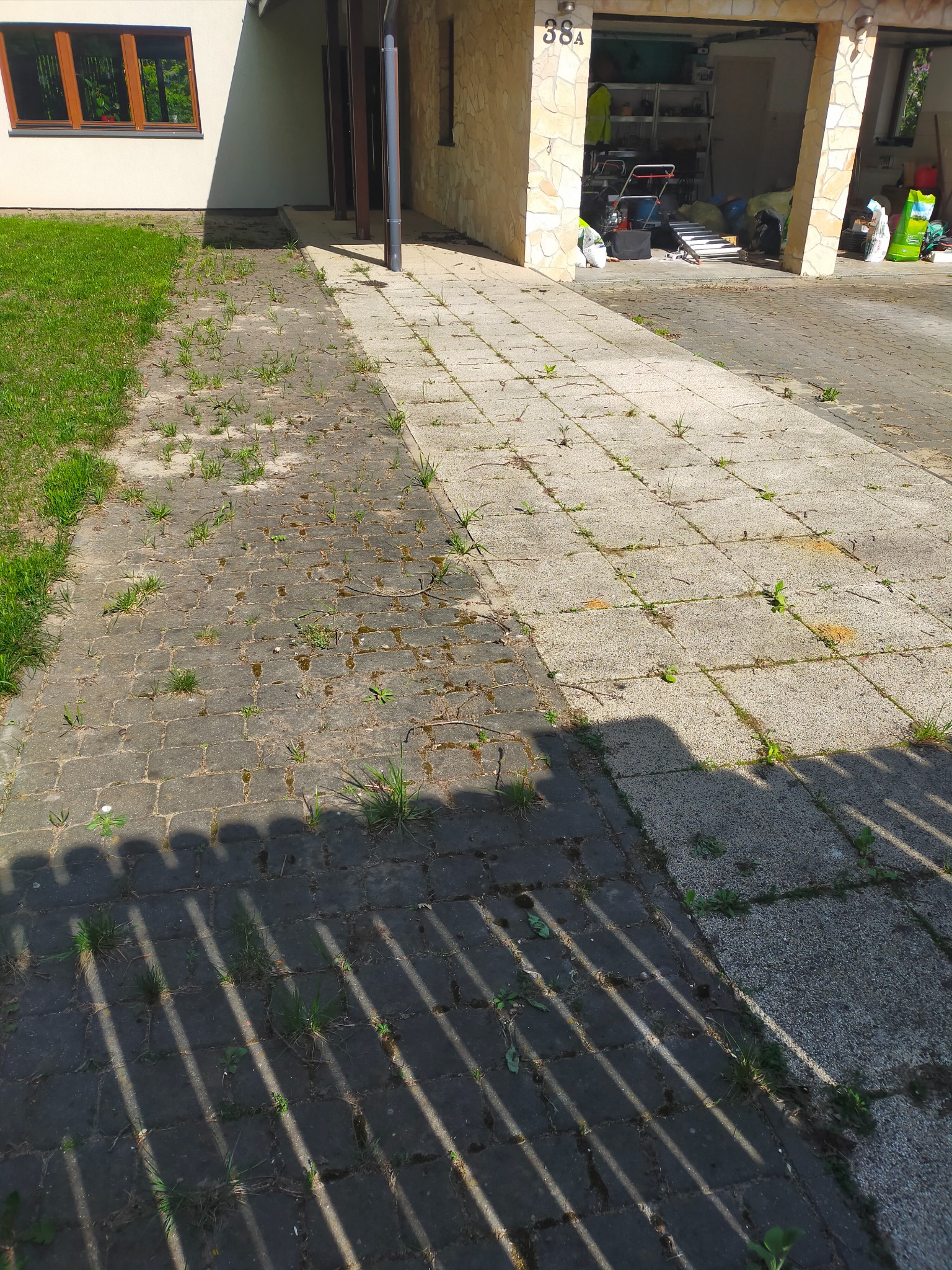 Porównanie stanu kostki brukowej przed i po czyszczeniu, widoczne różnice w kolorze i usunięte chwasty, cień płotu na dolnym planie.