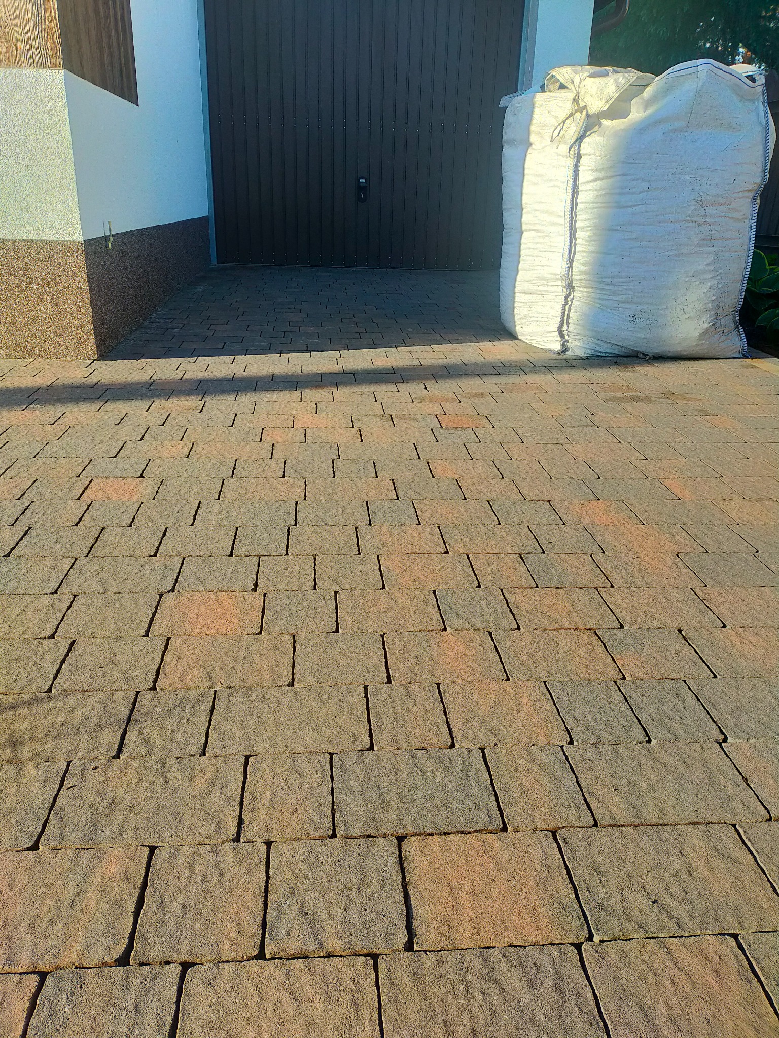 Ułożona kostka brukowa w odcieniach brązu i szarości, z widocznym workiem z piaskiem obok garażu, rzucająca cień na nawierzchnię.