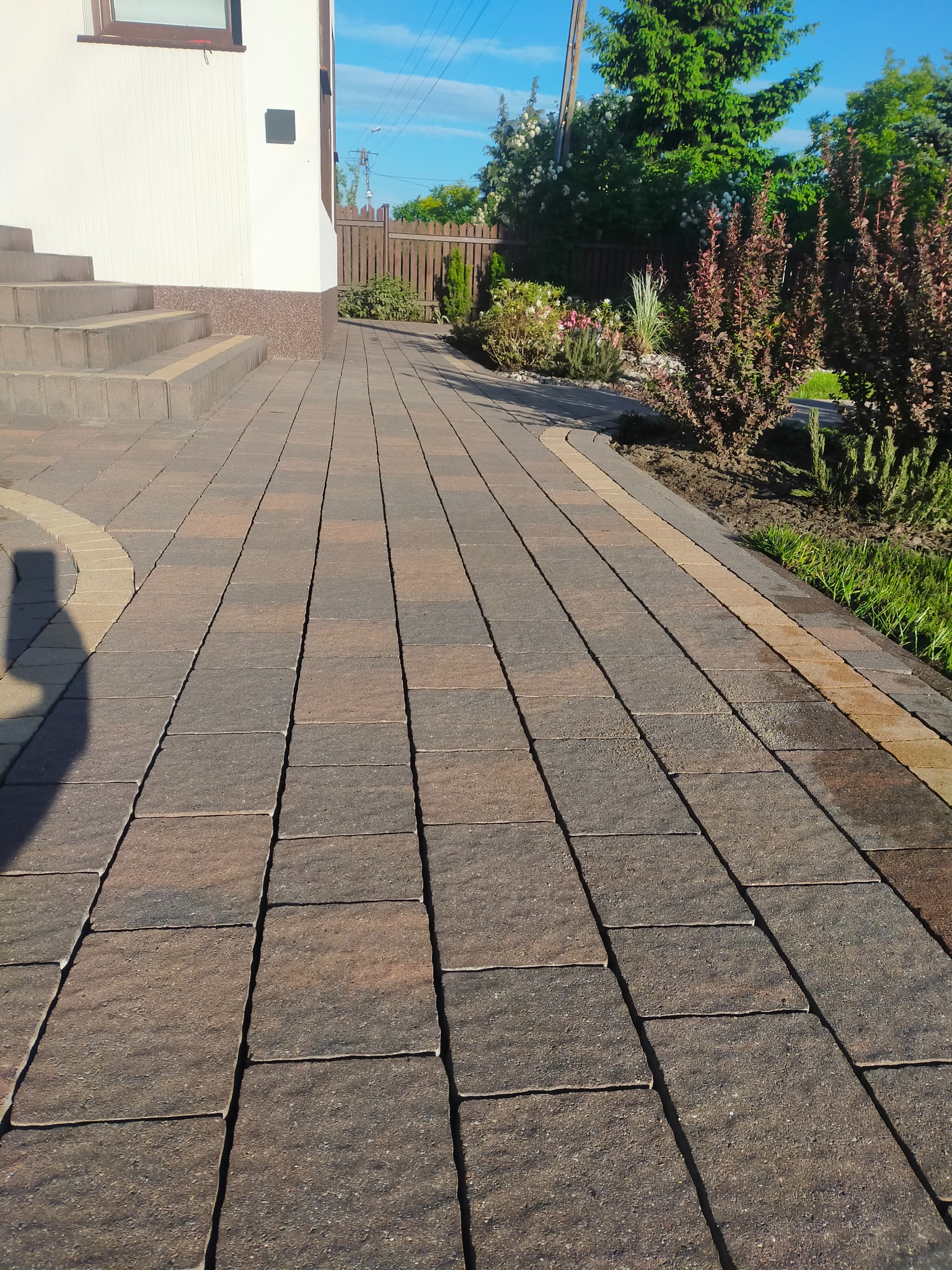 Ułożona, brązowo-szara kostka brukowa prowadząca od schodów domu w kierunku ogrodu z roślinami ozdobnymi i drewnianym płotem.