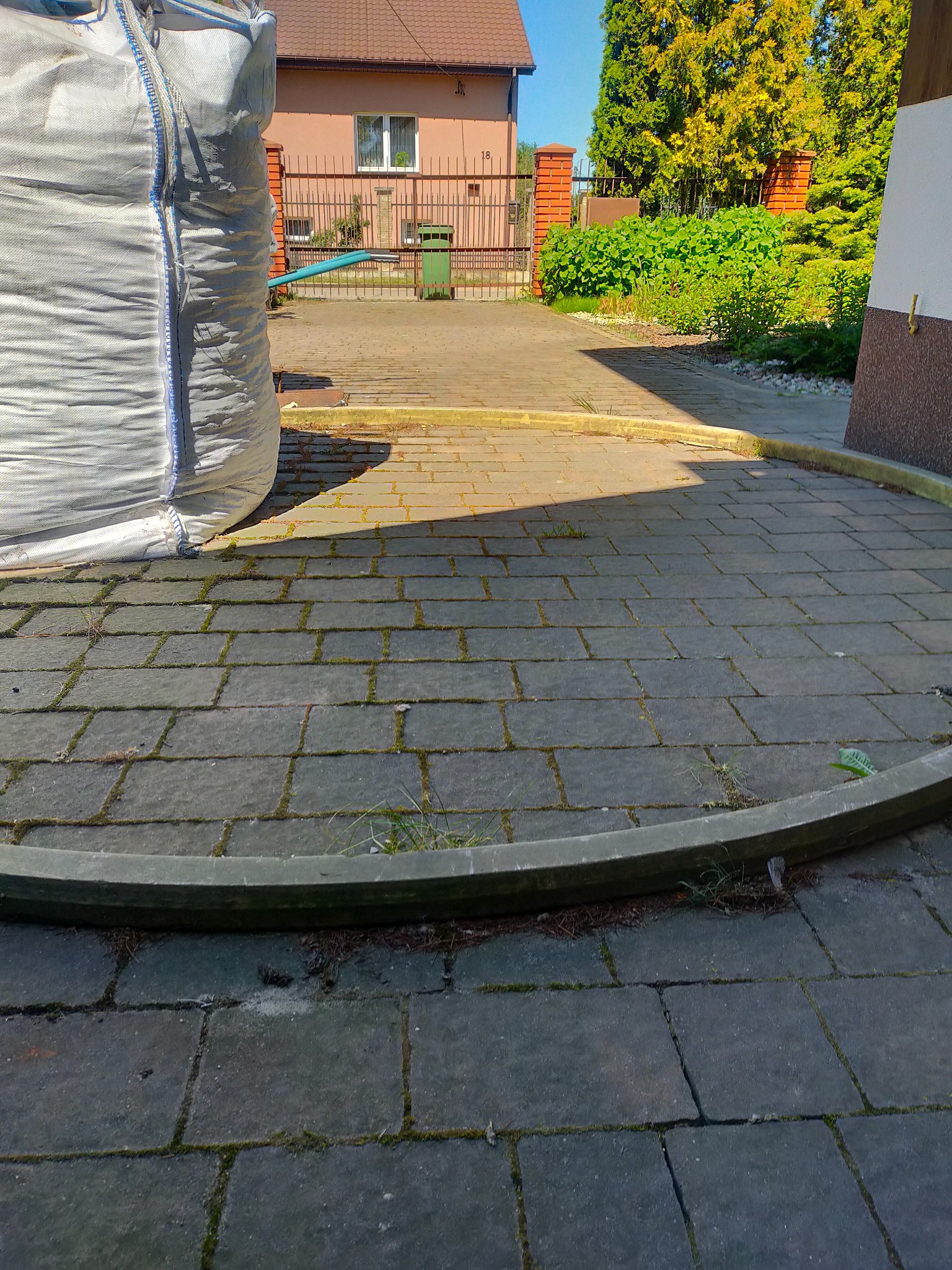 Szara kostka brukowa z mchem w spoinach, częściowo oświetlona słońcem, obok duży biały worek typu Big Bag i zielony trawnik w tle.