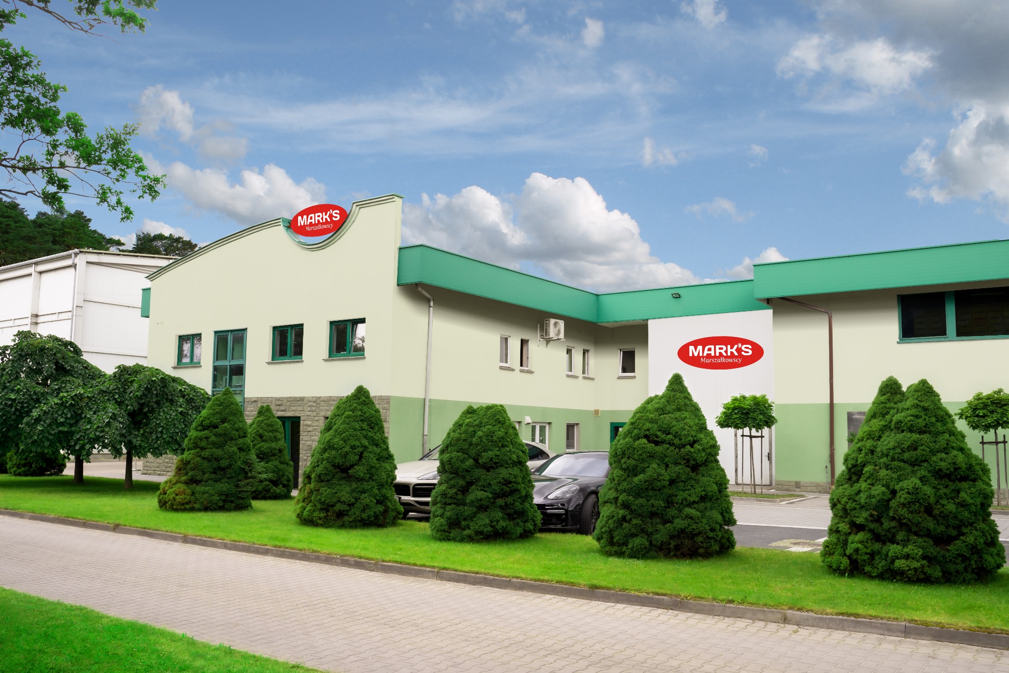 Budynek firmy Mark's Marszałkowscy z logo na elewacji, otoczony zielenią i zaparkowanymi samochodami.