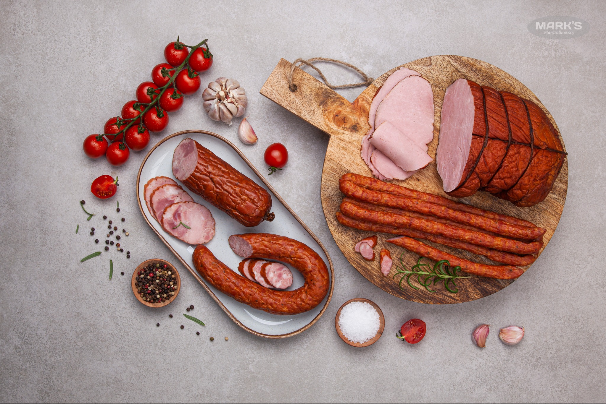 Kompozycja wędlin i dodatków: wędzona szynka w siatce, kabanosy, kiełbasa wędzona w plasterkach na talerzu, pomidorki cherry, czosnek, sól i pieprz na szarym tle.