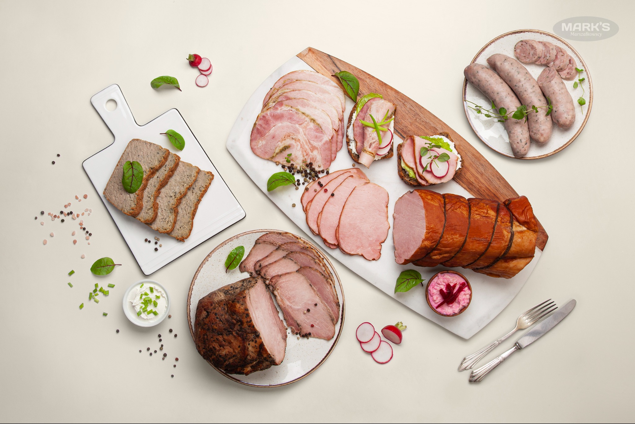 Kompozycja różnorodnych wędlin, pasztetu i kanapek z dodatkami, ułożonych na deskach i talerzach, z widocznymi przyprawami i ziołami, tworząca apetyczną prezentację.