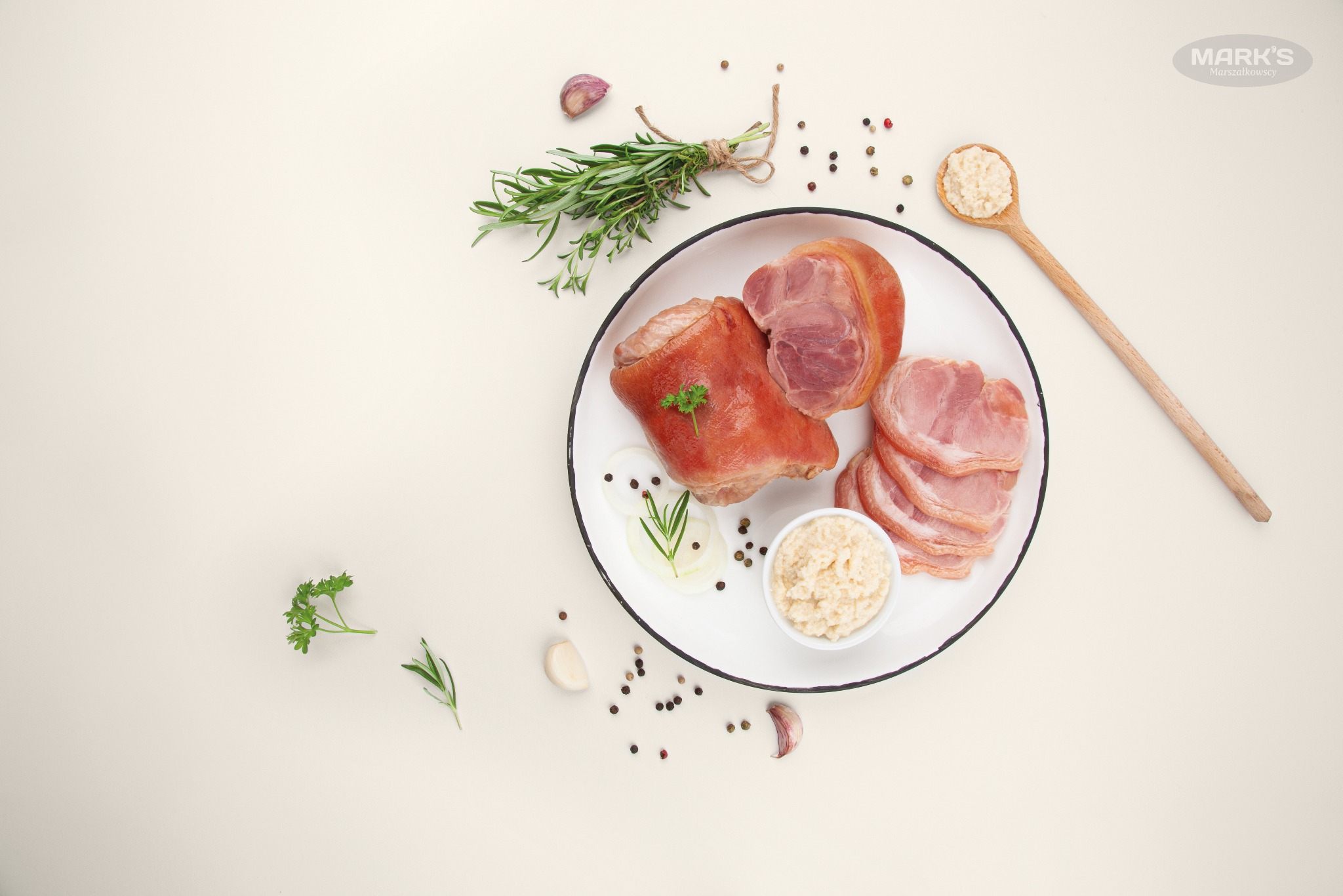 Szynka gotowana w całości i w plastrach na białym talerzu z chrzanem, cebulą, czosnkiem, rozmarynem i ziarnami pieprzu na jasnym tle, drewniana łyżka z chrzanem obok.