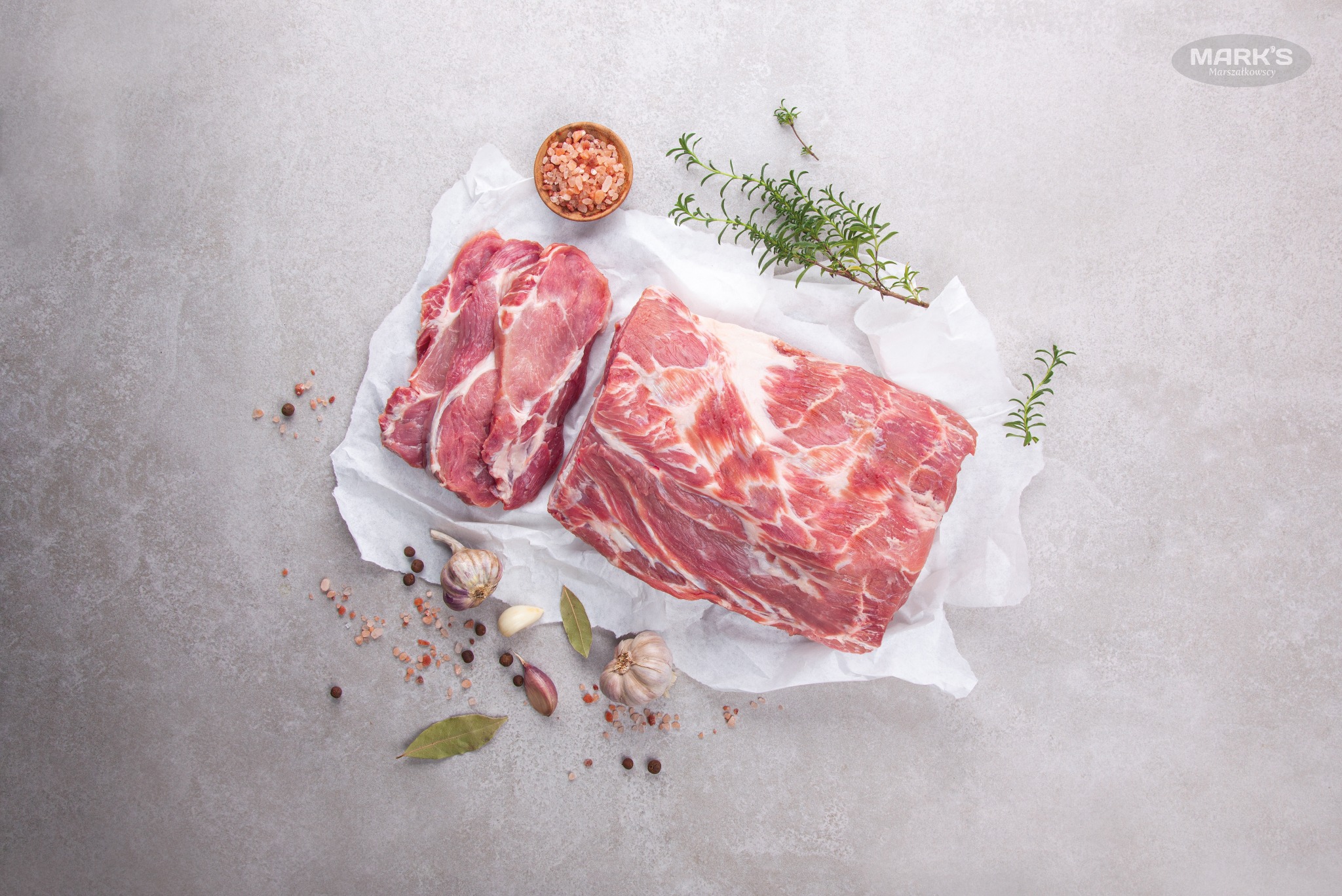 Surowe kawałki karkówki wieprzowej na białym papierze pergaminowym, obok przyprawy: sól himalajska w drewnianej miseczce, czosnek, ziele angielskie, liście laurowe i gałązka rozmarynu, całość...