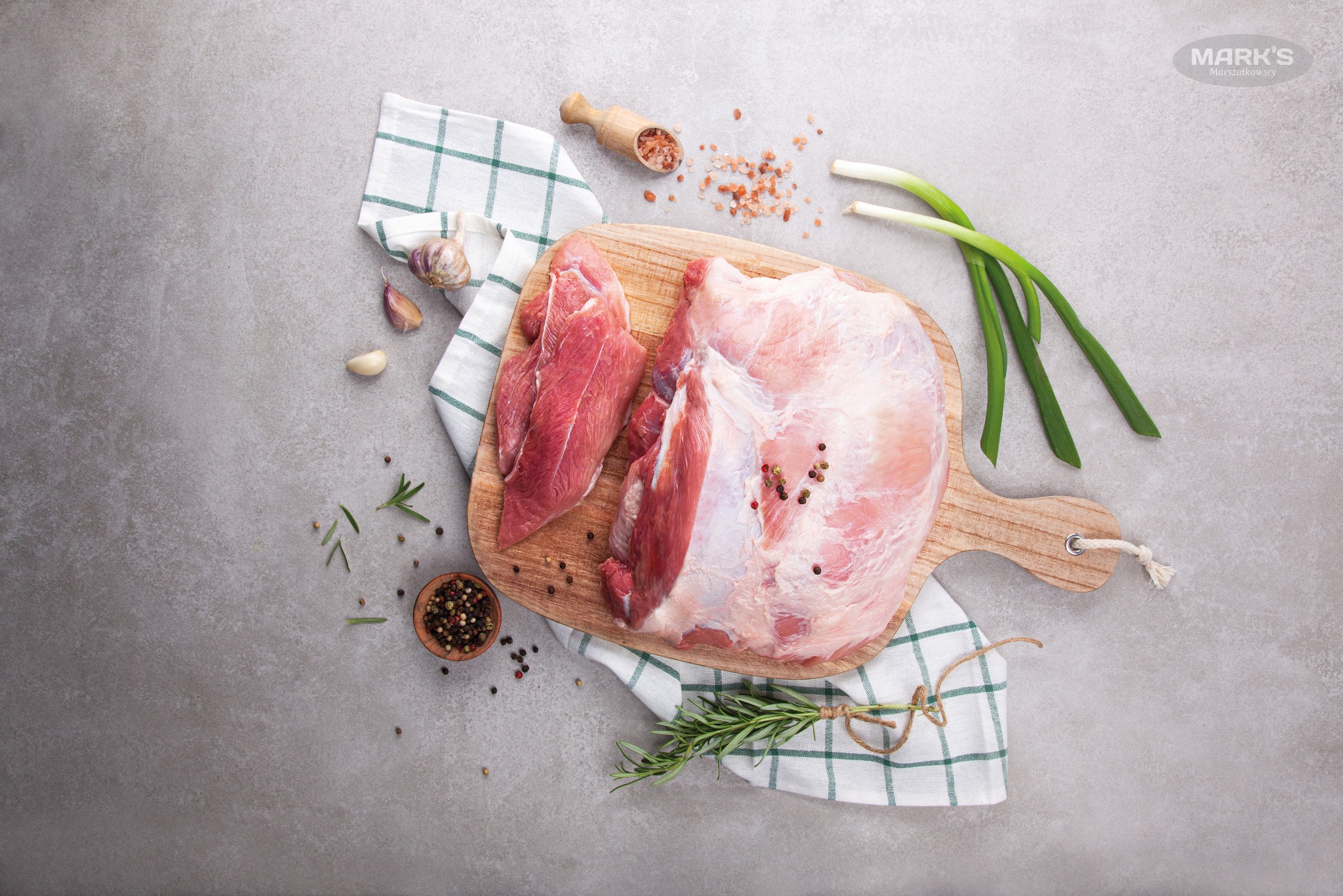 Surowe mięso wieprzowe i wołowe na drewnianej desce do krojenia, otoczone przyprawami, czosnkiem, cebulą dymką i ściereczką w kratę na szarym tle.