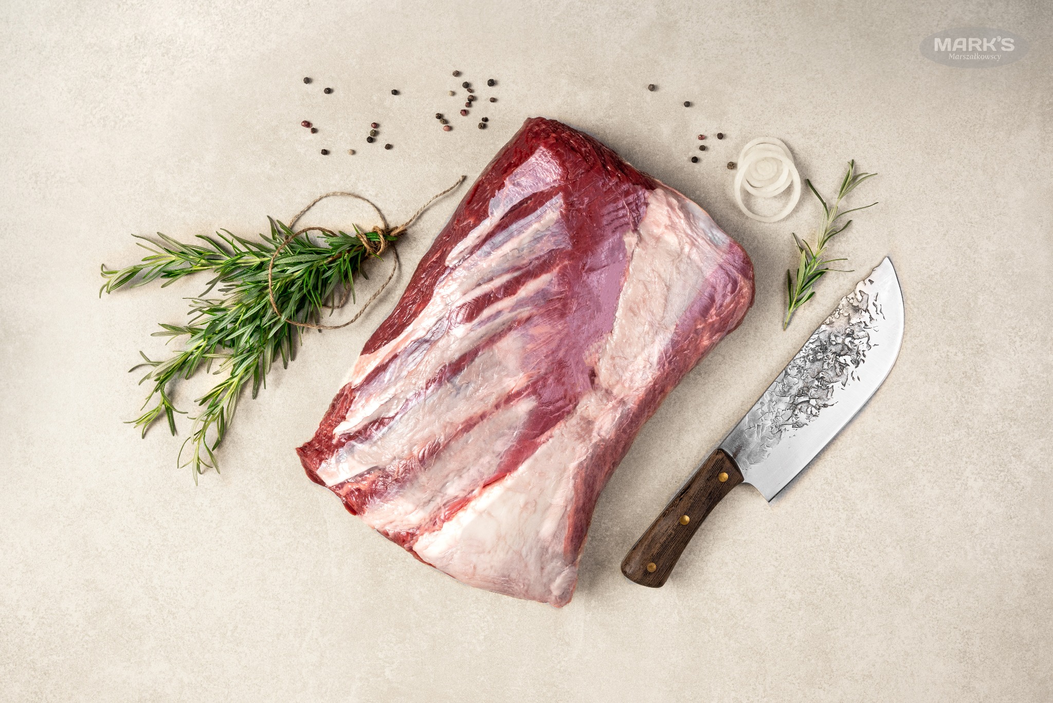 Surowy kawał wołowiny z żebrami, obok pęczek rozmarynu związany sznurkiem, ziarna pieprzu, krążki cebuli i nóż z drewnianą rękojeścią na jasnym tle.