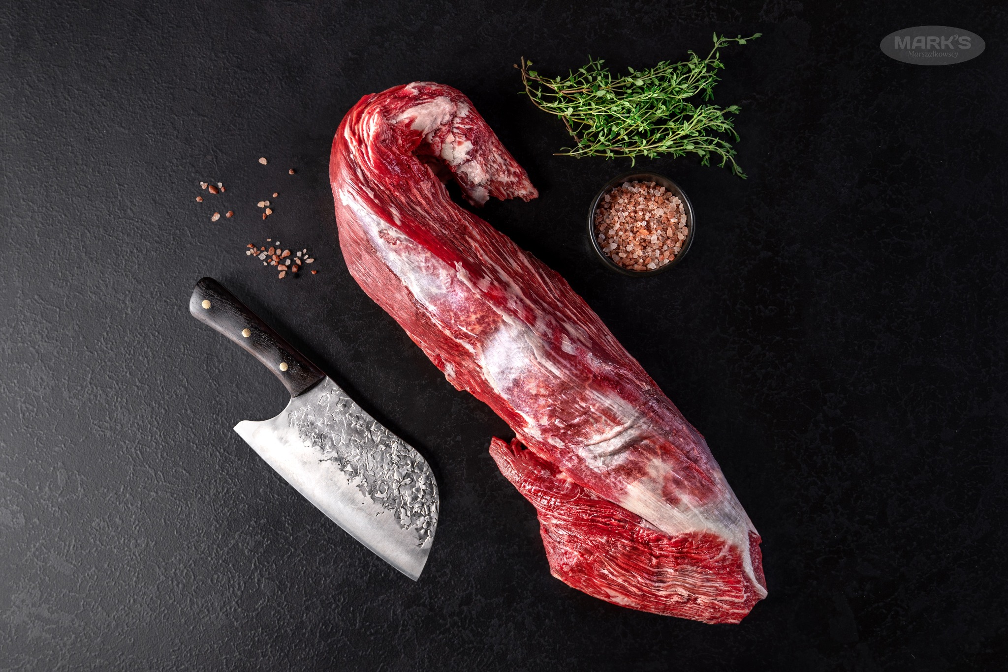 Surowy rostbef wołowy z marmurkowym wzorem, nóż rzeźniczy z kutym ostrzem, gałązki tymianku i różowa sól himalajska na czarnym tle.