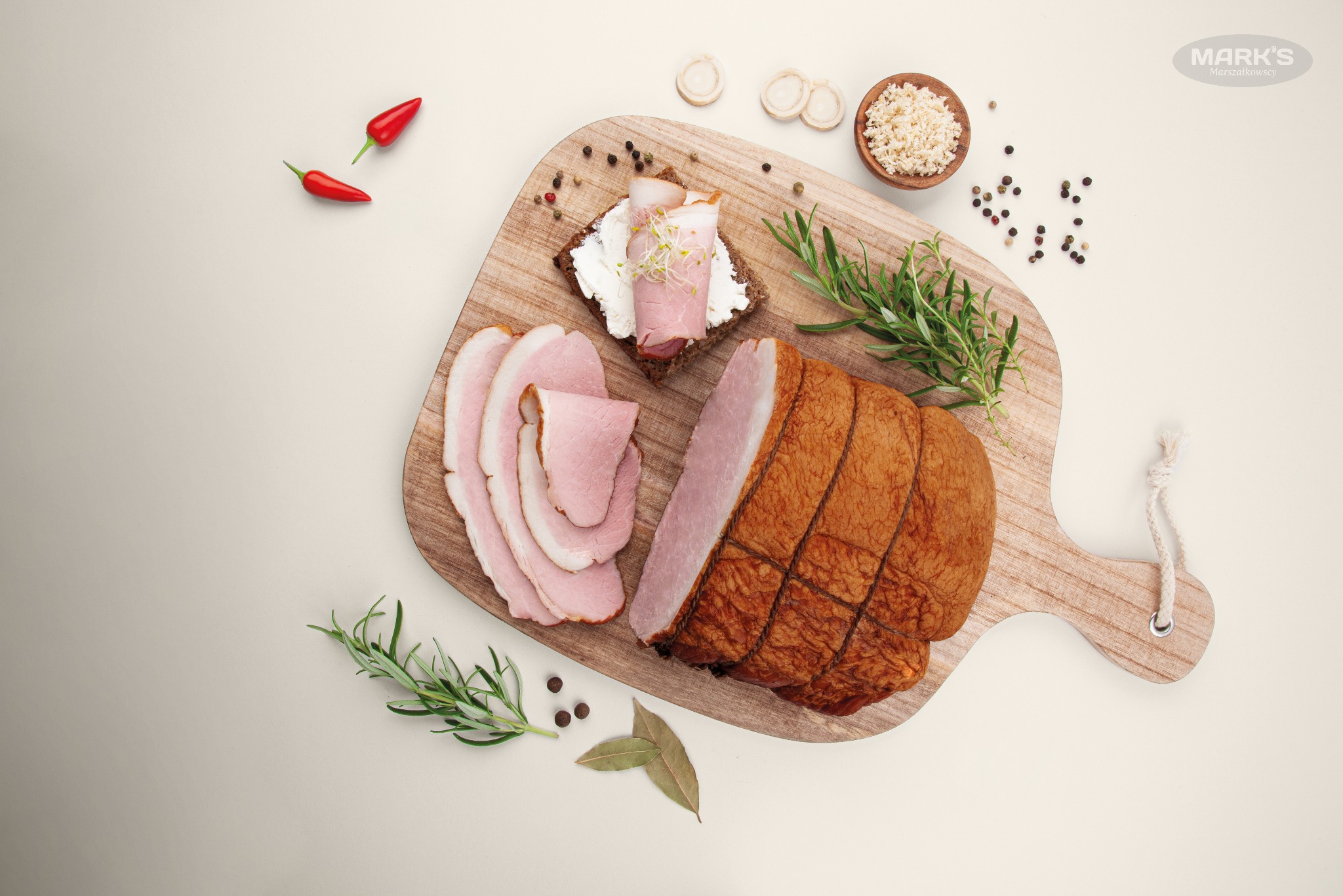 Deska do krojenia z wędlinami, kromką chleba z kremowym serem i kiełkami, przyprawami i ziołami, widok z góry, ostre papryczki w tle.