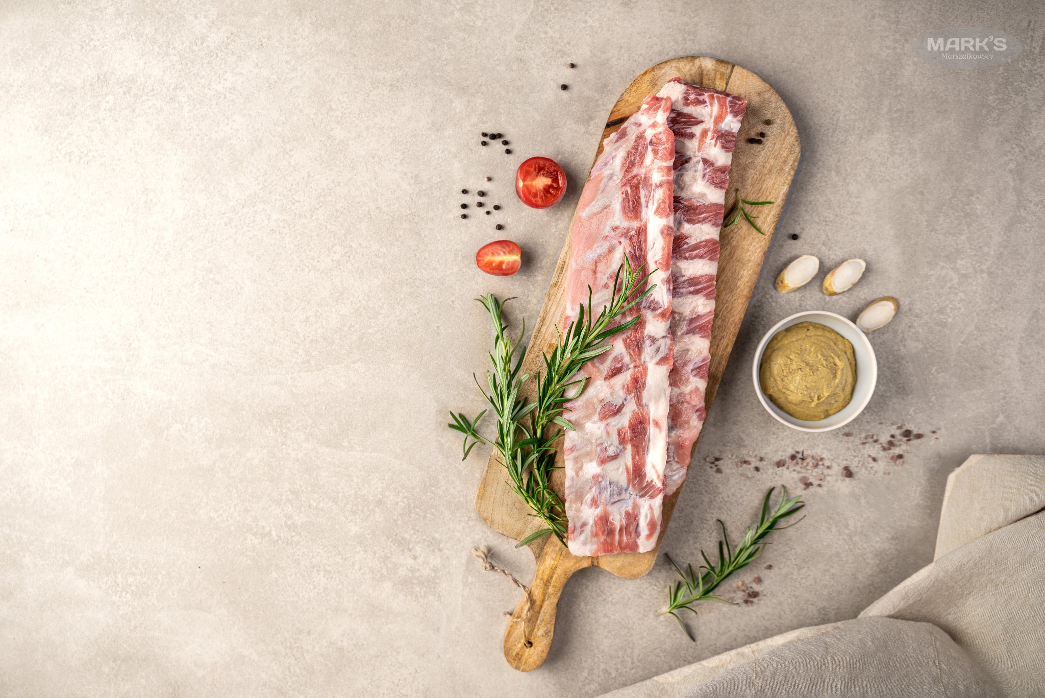 Surowe żeberka wieprzowe na drewnianej desce do krojenia, z rozmarynem, połówkami pomidora, ziarnami pieprzu i sosem musztardowym na szarym tle.