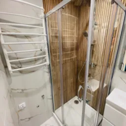 Nowoczesna łazienka z kabiną prysznicową w rogu, wykończona drewnianą okładziną. Białe marmurowe płytki i grzejnik drabinkowy. Widok na umywalkę i białą podłogę.