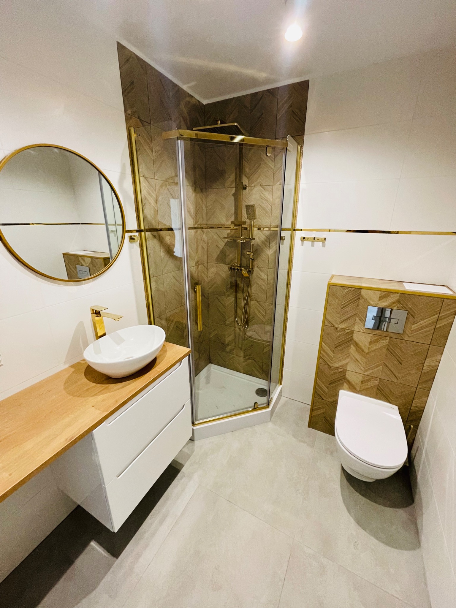 Nowoczesna łazienka z kabiną prysznicową w złotej ramie, umywalką nablatową na drewnianym blacie i toaletą podwieszaną z zabudową w jodełkę, uzupełniona okrągłym lustrem.