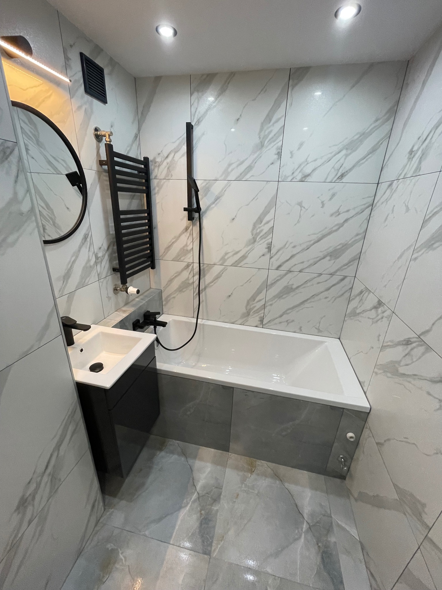Nowoczesna łazienka z marmurowymi płytkami, czarną armaturą, grzejnikiem drabinkowym i okrągłym lustrem, oświetlona punktowymi lampami sufitowymi.