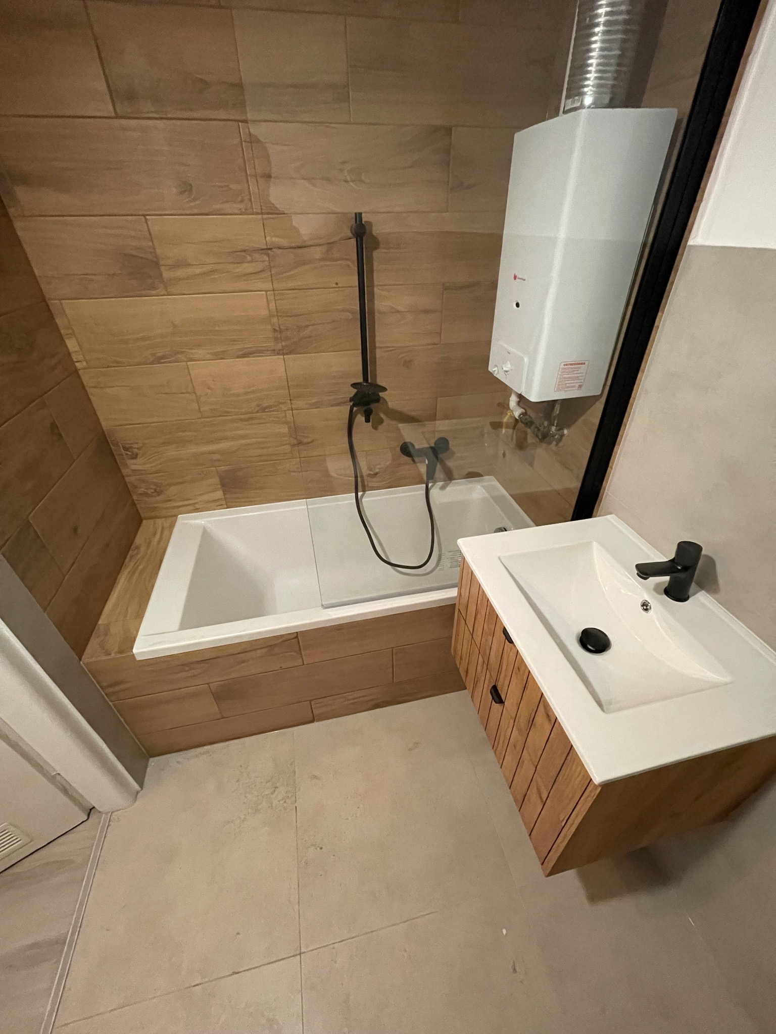 Nowoczesna łazienka z wanną, umywalką z szafką z drewnianymi lamelami, brązowymi płytkami imitującymi drewno na ścianach i białym bojlerem.