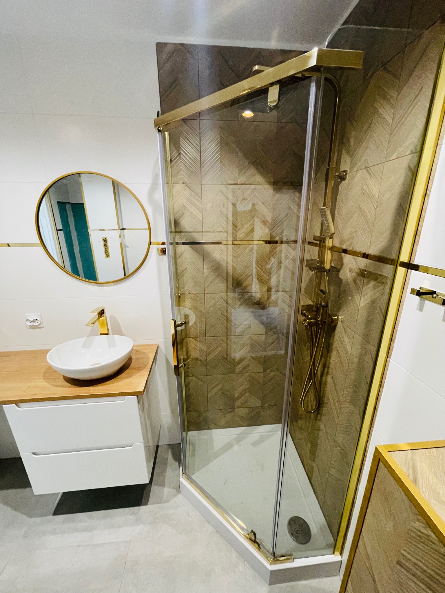 Nowoczesna łazienka z kabiną prysznicową narożną ze złotymi profilami, umywalką nablatową na szafce z drewnianym blatem i okrągłym lustrem w złotej ramie.