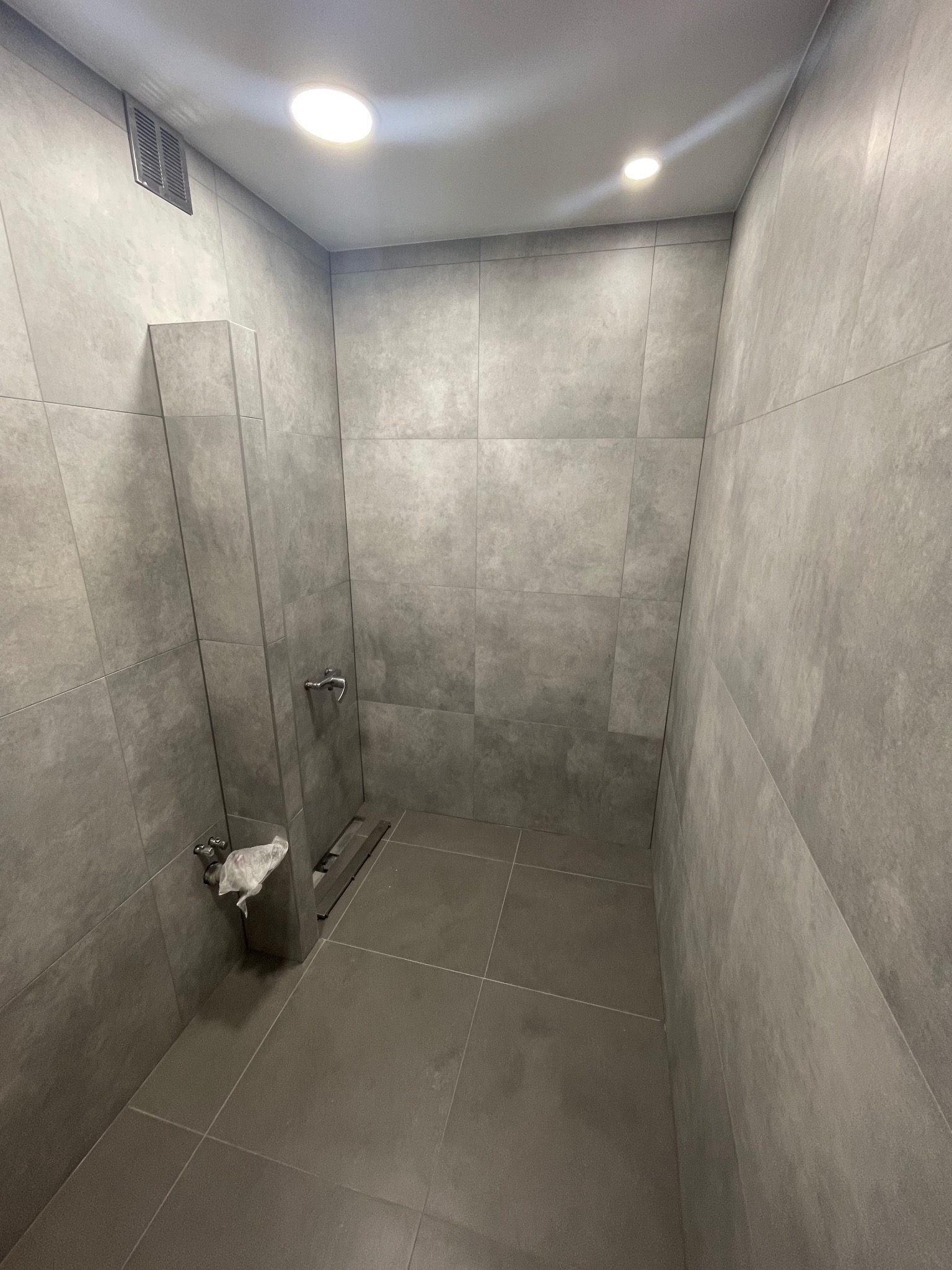 Wnętrze kabiny prysznicowej wyłożonej szarymi płytkami imitującymi beton, z widocznym odpływem liniowym, baterią prysznicową i fragmentem instalacji wodnej zabezpieczonej folią.