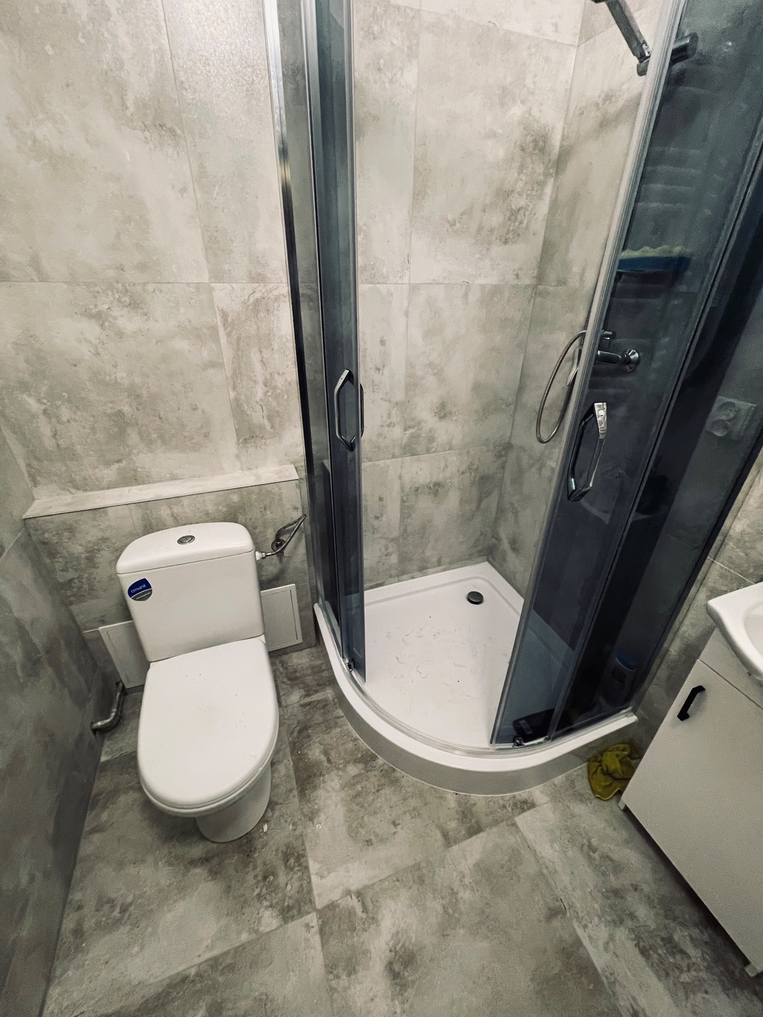 Nowoczesna łazienka z szarymi płytkami imitującymi beton, kabina prysznicowa z chromowanymi profilami i biała toaleta z widocznym logo producenta.