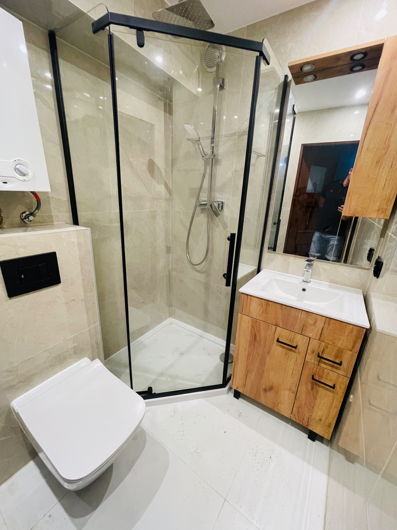 Nowoczesna łazienka po remoncie z kabiną prysznicową typu walk-in z czarnymi profilami, wiszącą toaletą, umywalką z szafką z frontami imitującymi drewno i dużym lustrem.